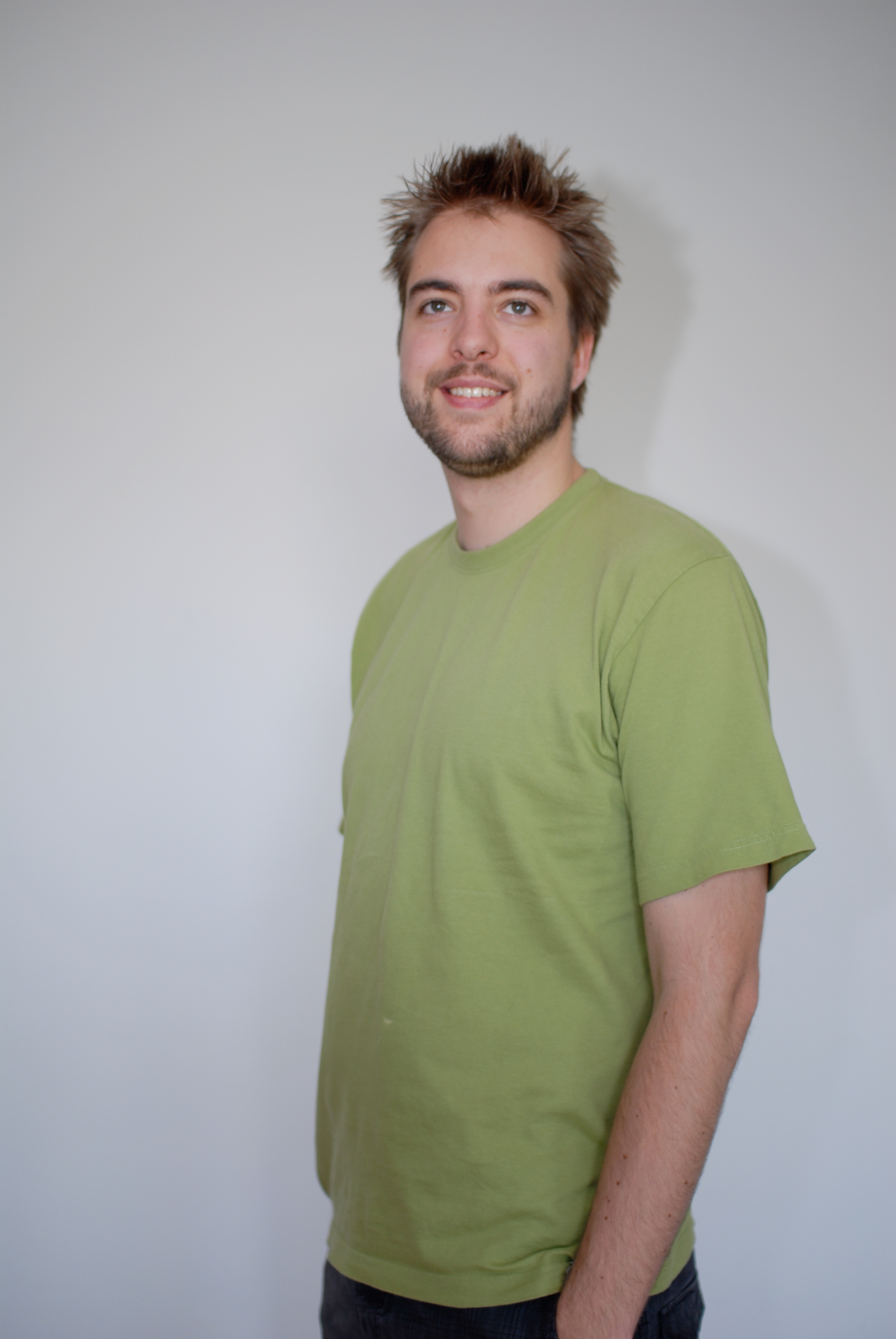 A man with short, spiky hair and a beard stands against a plain wall, wearing a green T-shirt and smiling.