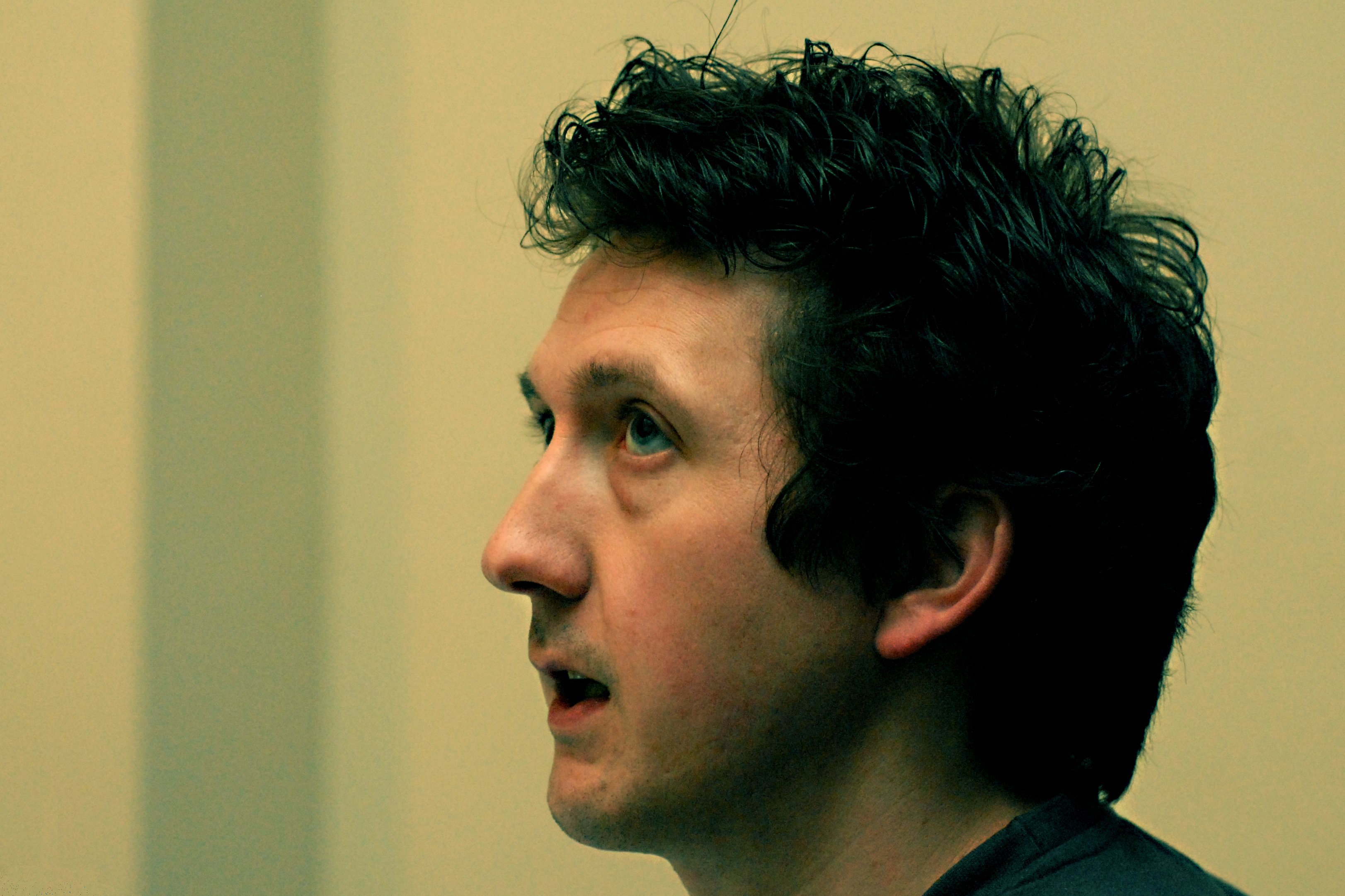A man with dark curly hair speaks while looking ahead in an indoor setting.