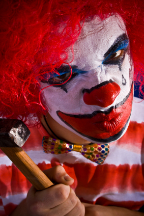A clown with red hair and face paint holds a hammer while staring intensely.