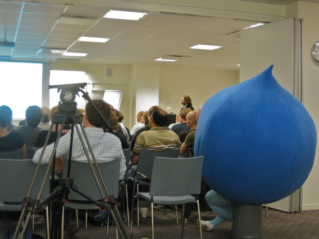 A person in a large blue Druplicon costume sits among an audience listening to a speaker in a conference room.