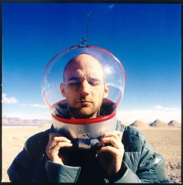 A man with closed eyes holds a clear astronaut-style helmet around his head in a desert landscape.