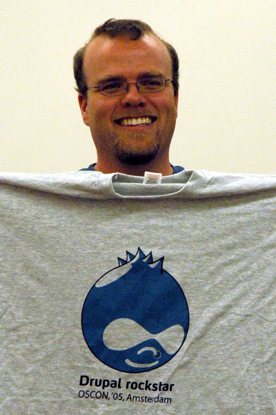 A smiling man holds up a gray T-shirt with a "Drupal rockstar" logo from OSCON 2005 in Amsterdam.