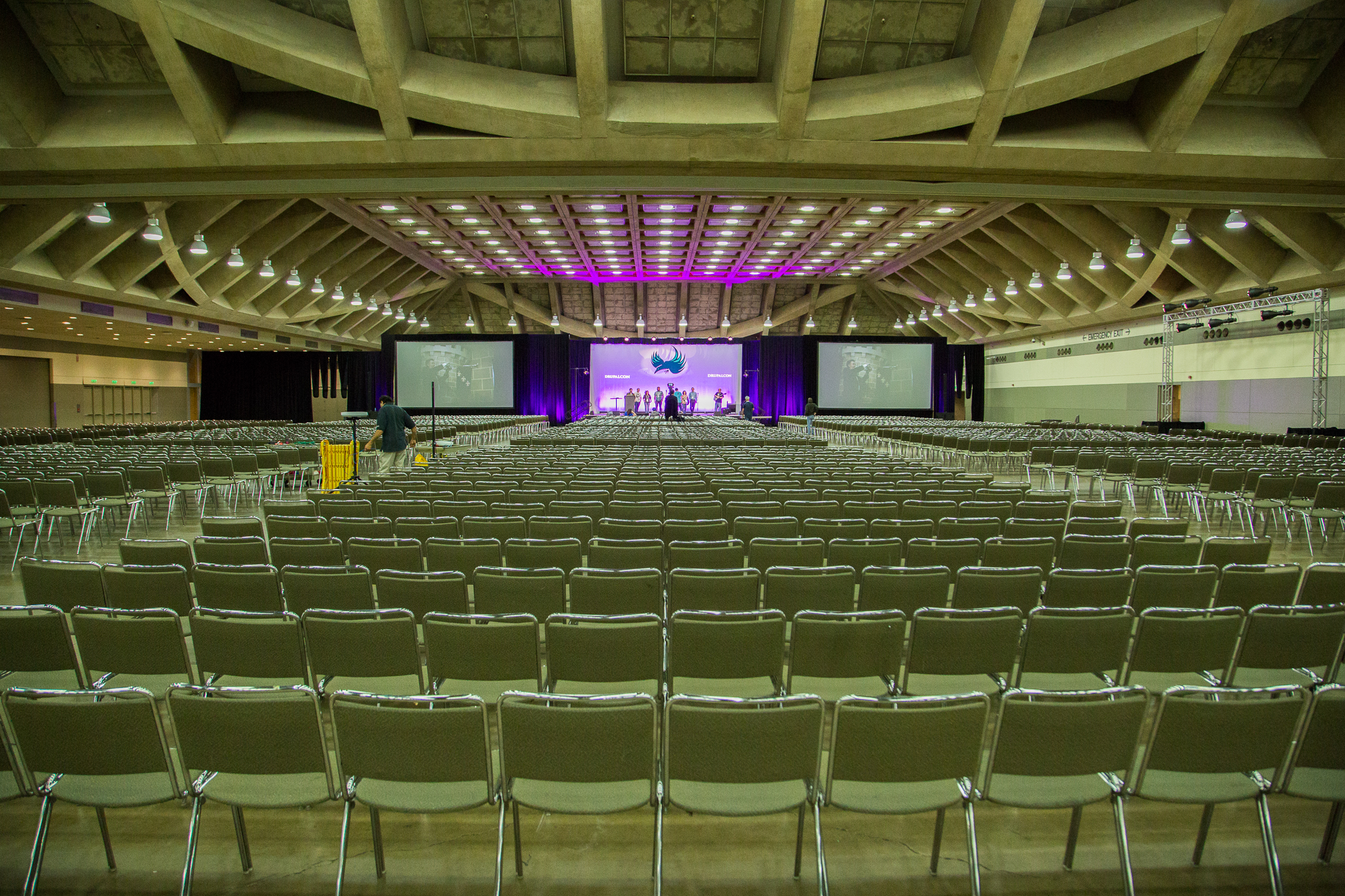 Large conference hall with rows of empty chairs facing a stage with screens and people preparing for an event.