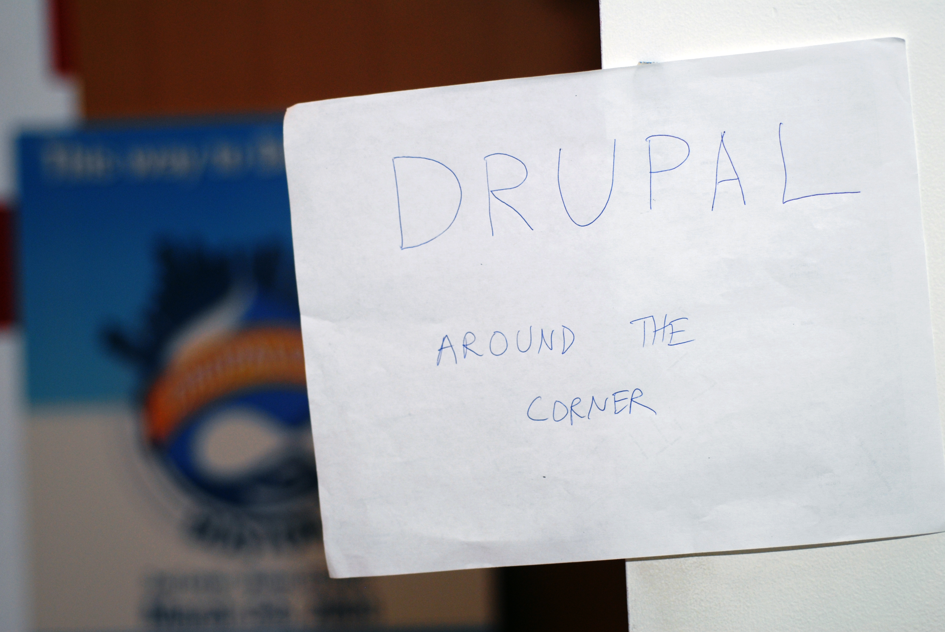 A handwritten sign on a wall reads "Drupal around the corner", directing attendees to a Drupal-related event nearby.