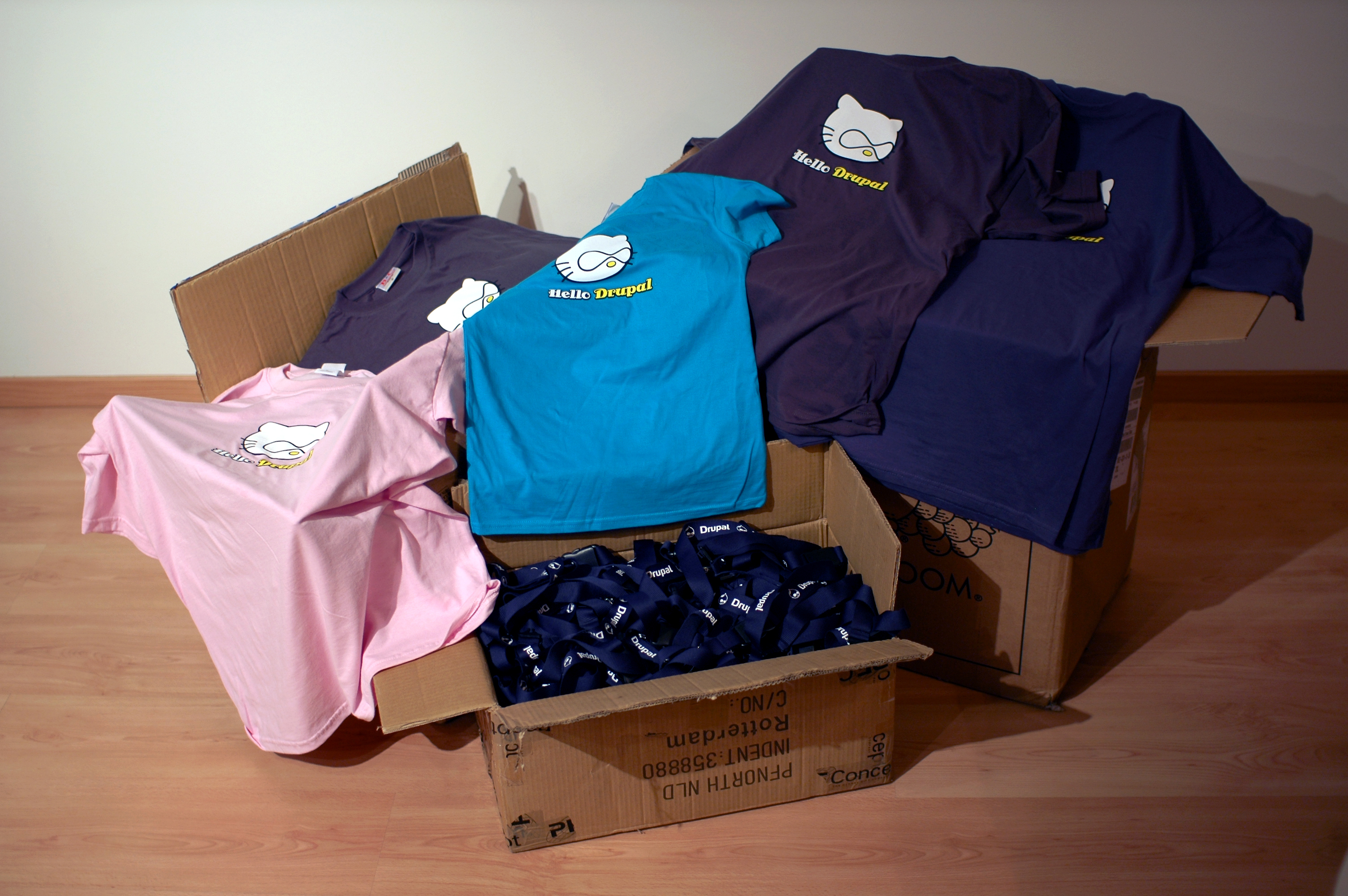Boxes filled with Drupal-themed t-shirts and lanyards are open and displayed on a wooden floor.