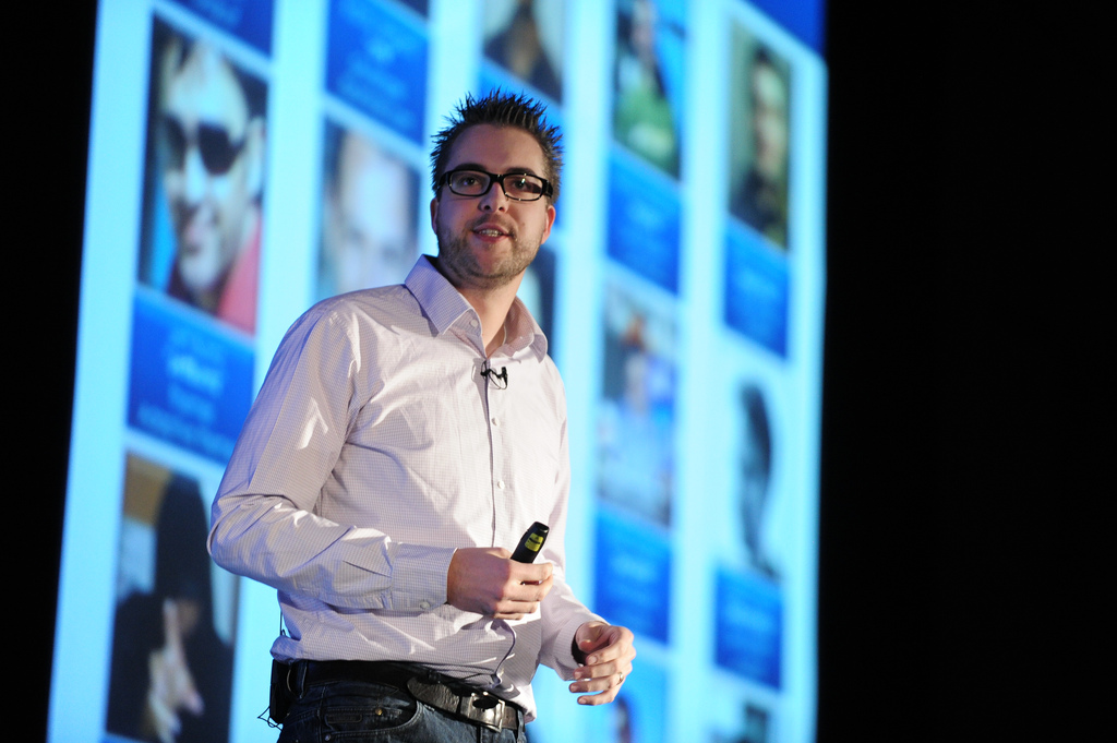 A speaker in a light-colored shirt gives a keynote presentation while holding a remote, with a screen behind him.