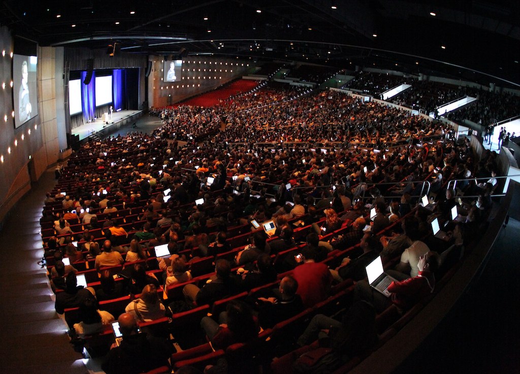 A large audience watches a keynote speaker on stage at DrupalCon Denver 2012, with many using laptops.