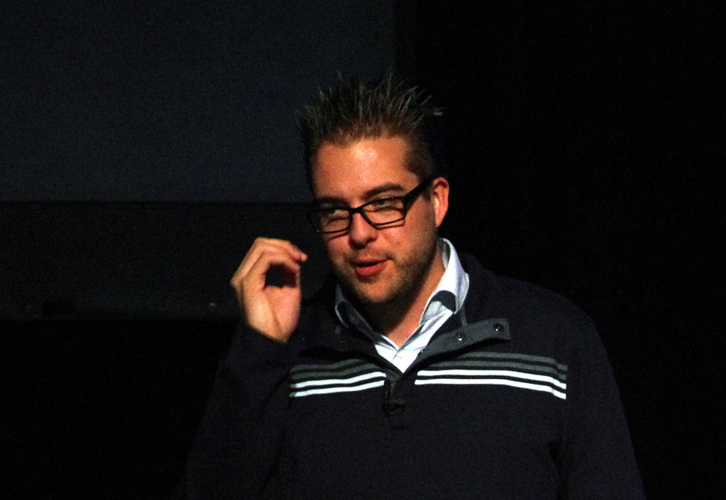 A speaker wearing glasses and a dark sweater presents on stage during the DrupalCon Denver 2012 keynote.