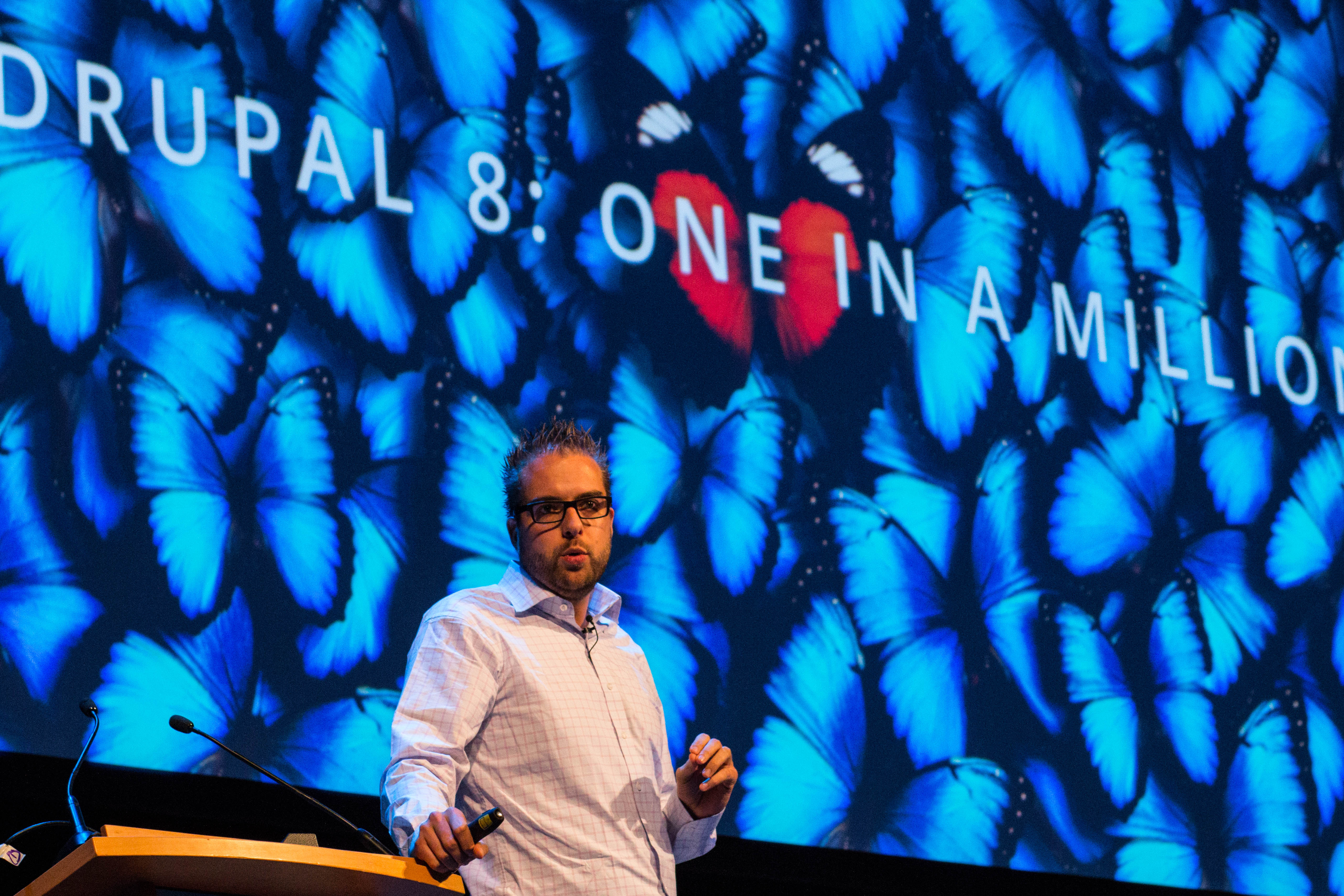 A speaker presents on stage at DrupalCon Dublin 2016, standing in front of a large screen with blue butterflies.