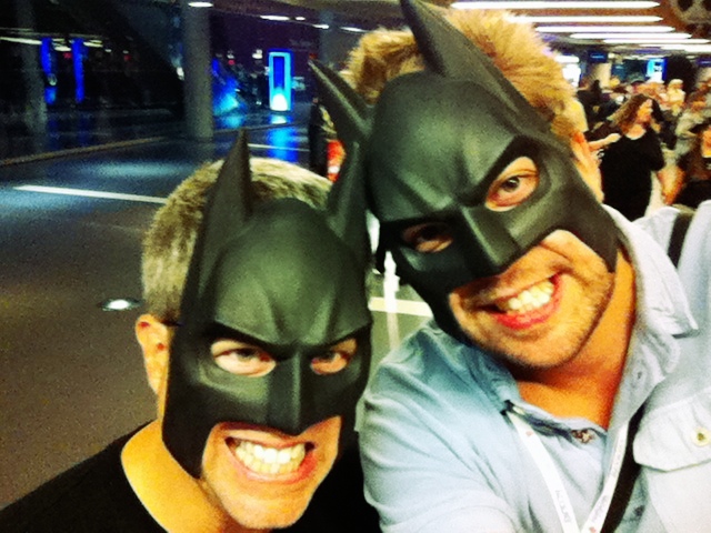 Two people wearing Batman masks smile and pose for a selfie in a busy indoor setting.
