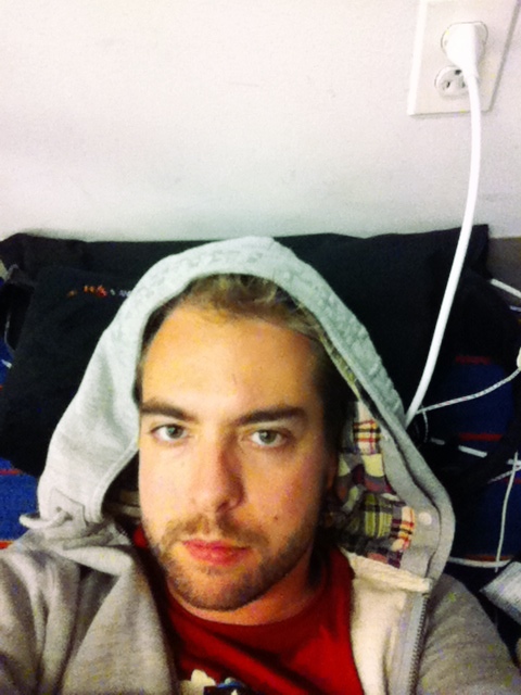 A person in a hoodie lies down near a wall with a plugged-in charger at JFK Airport.