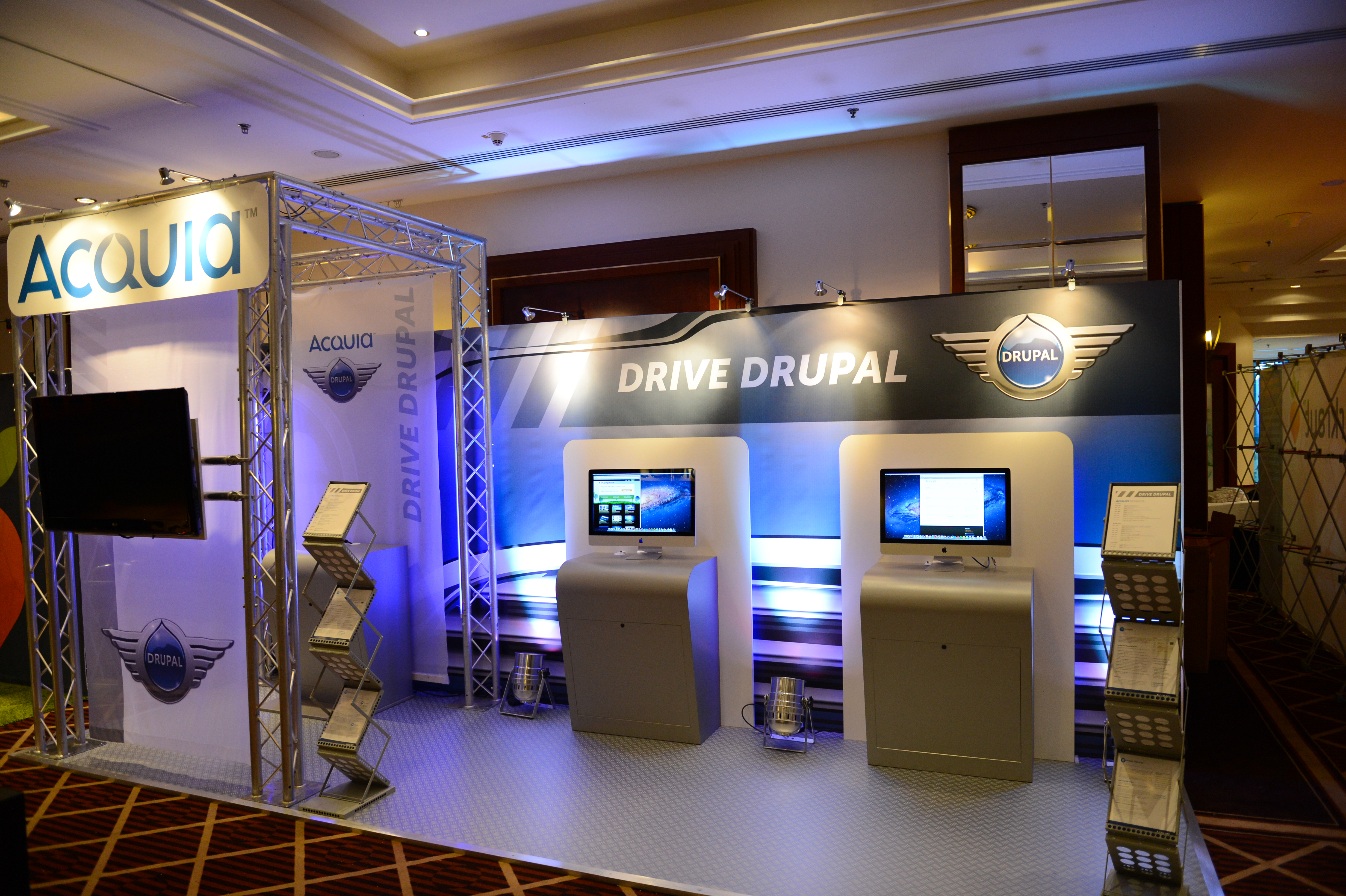 Acquia booth at DrupalCon Munich 2012 featuring two computer stations, promotional materials, and a "Drive Drupal" display.