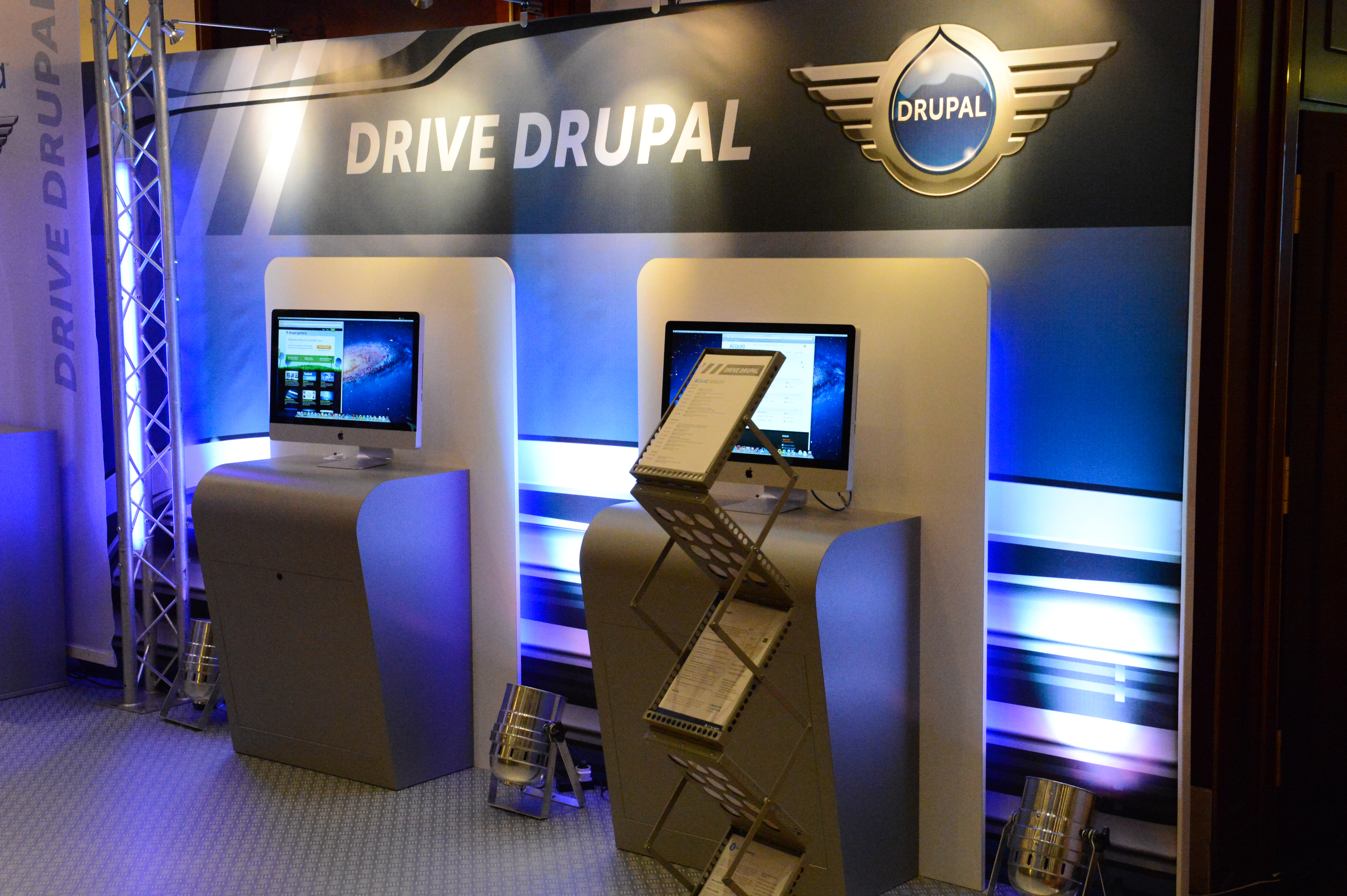 Acquia booth at DrupalCon Munich 2012 with two computer stations, promotional materials, and a "Drive Drupal" backdrop.
