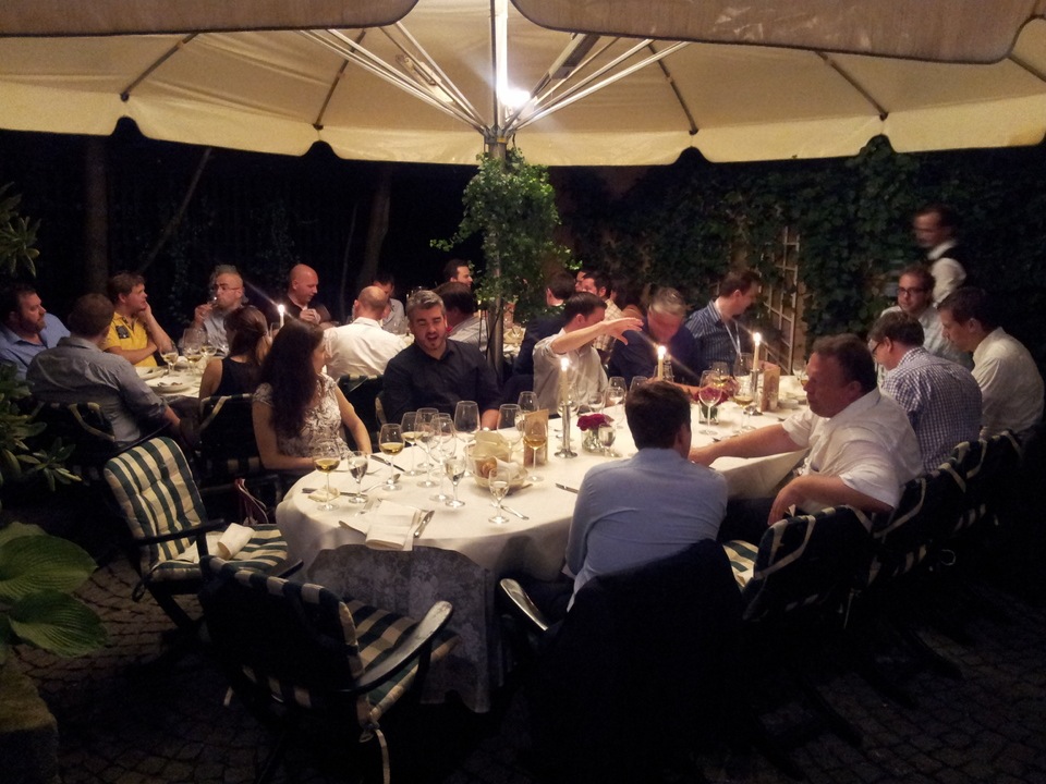 A group of people gathered around a table at an outdoor dinner, engaged in conversation and enjoying drinks.