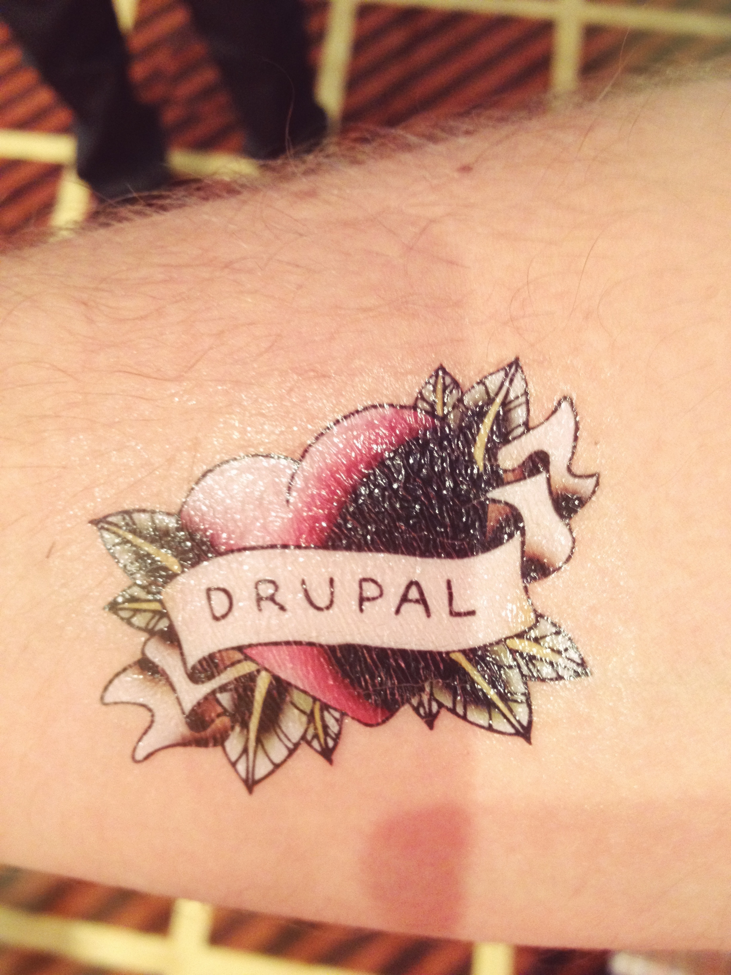 A close-up of a tattoo on an arm featuring the word "Drupal" on a banner with a heart design.