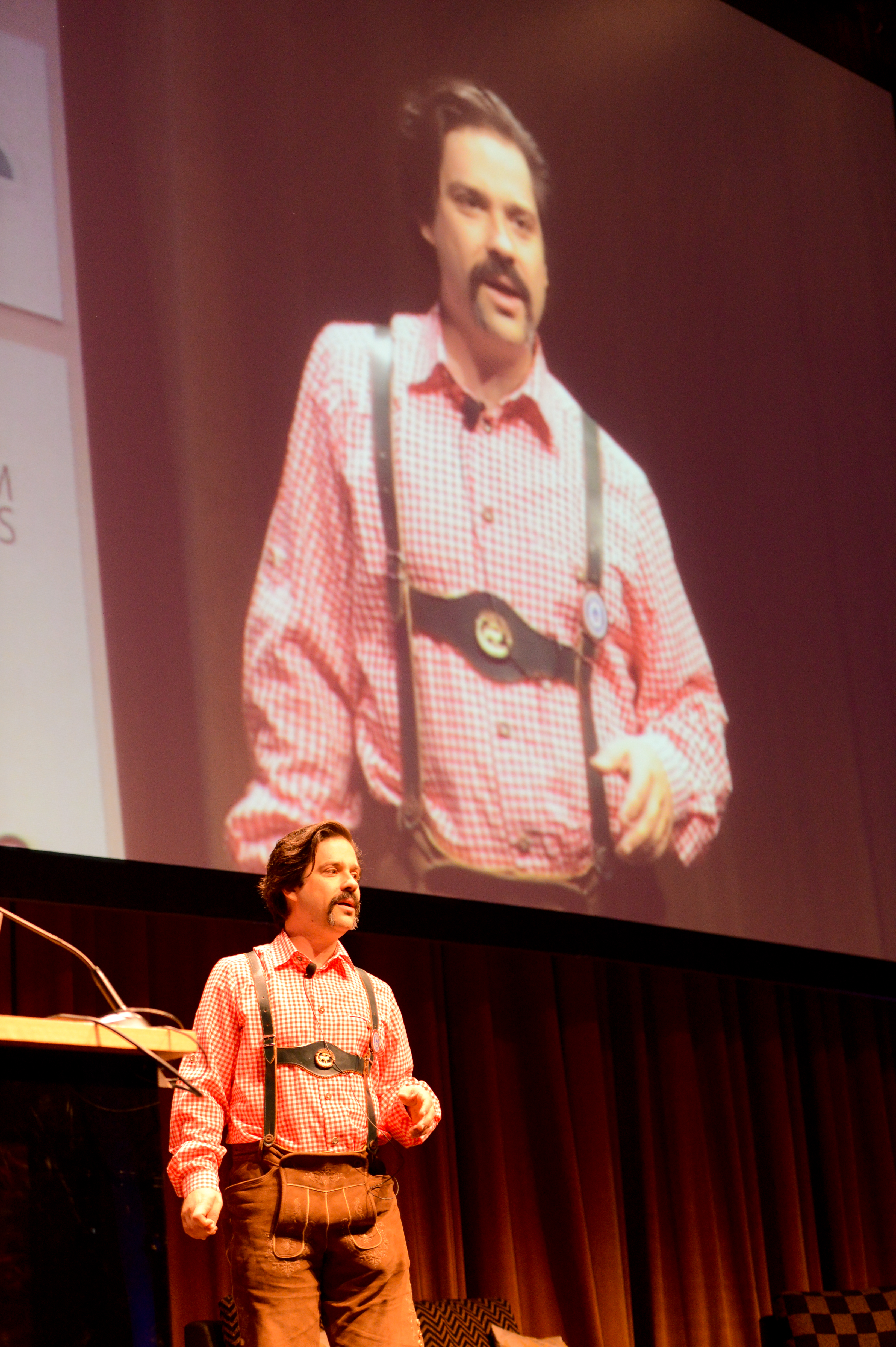 A speaker in traditional Bavarian attire presents on stage at DrupalCon Munich 2012, with a large screen behind him.