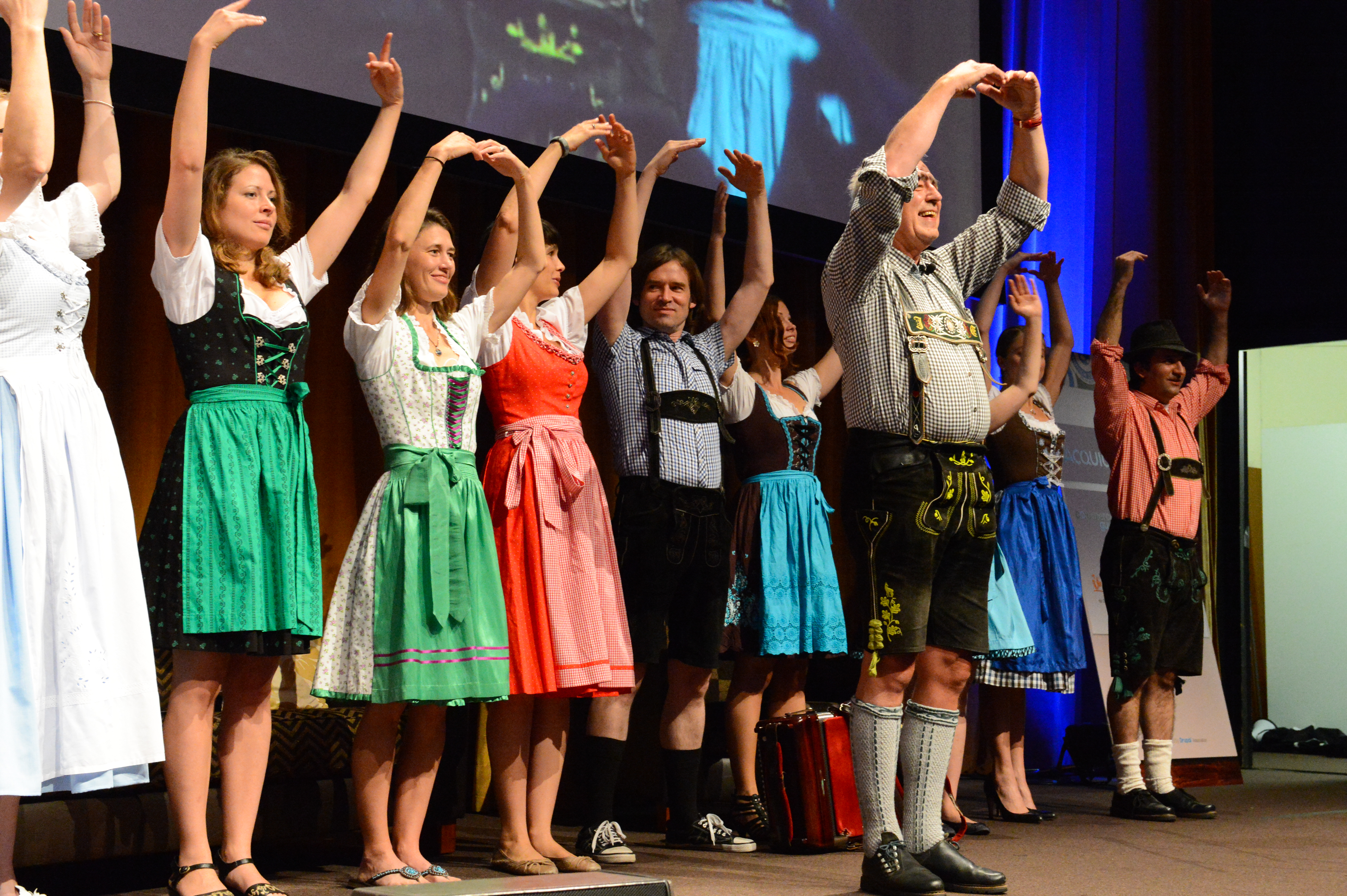 A group of people in traditional Bavarian clothing stand on stage with arms raised during an intro session.