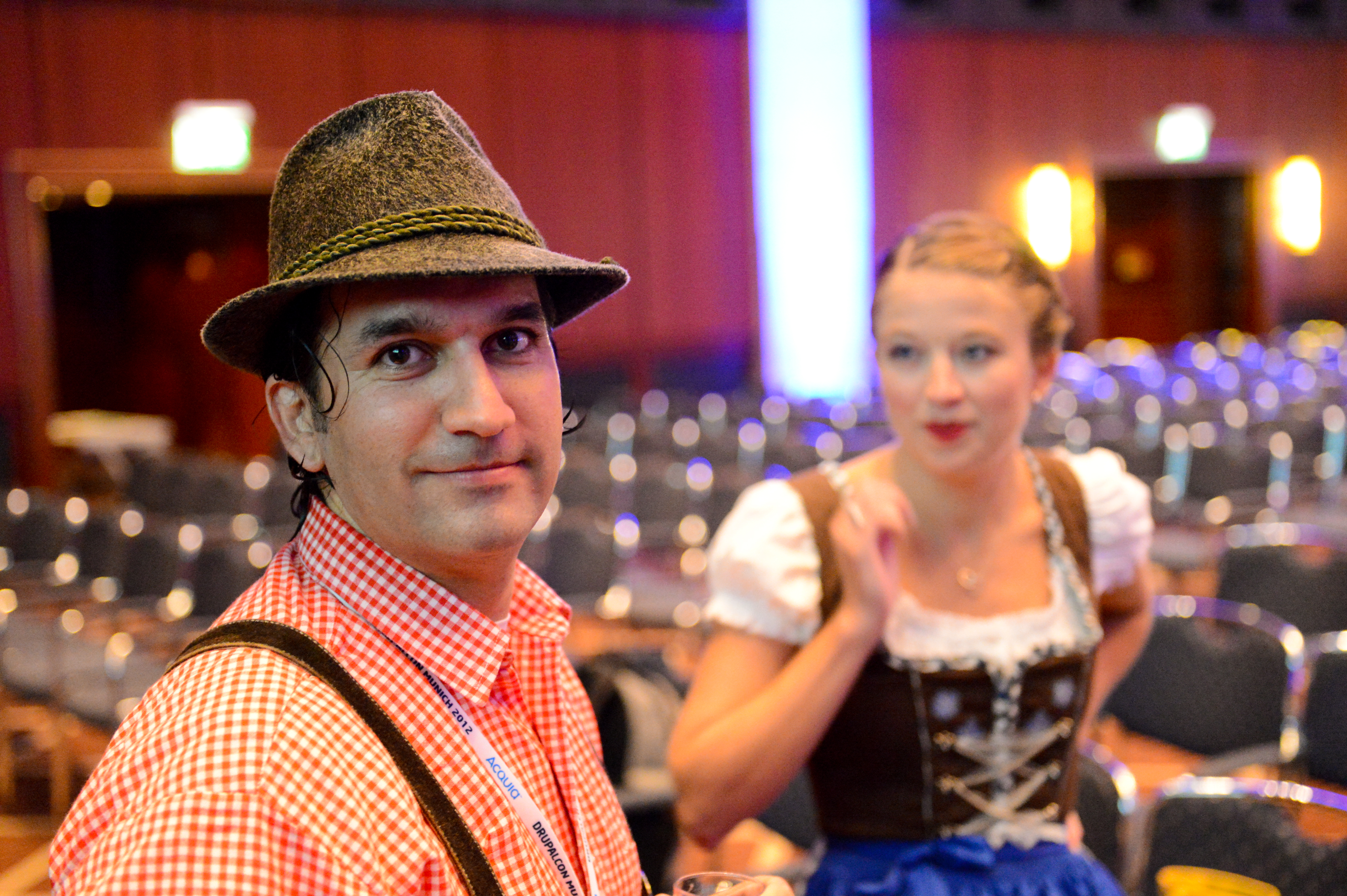 A man in traditional Bavarian clothing looks at the camera, while a woman in similar attire speaks in the background.