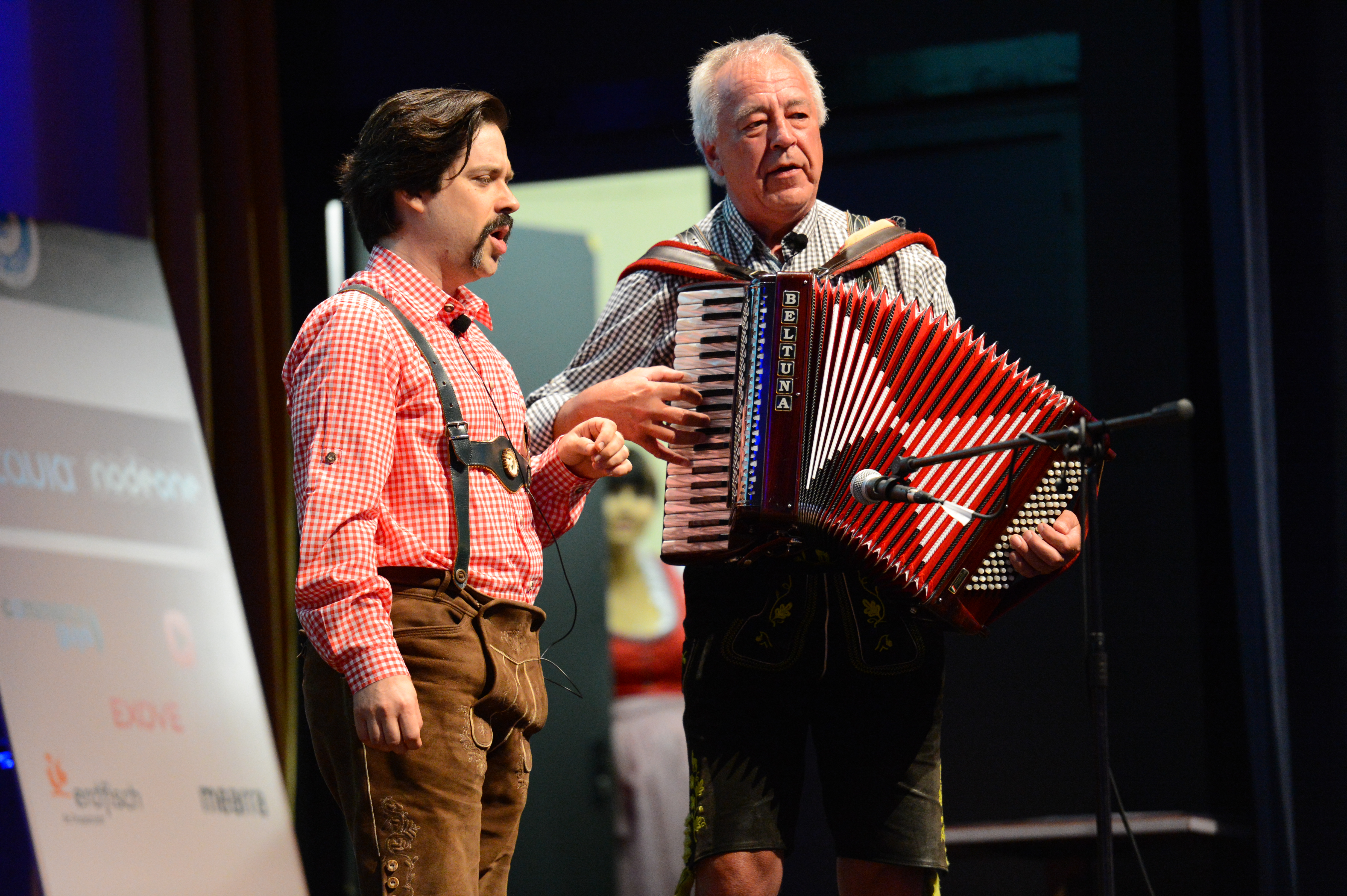Two men in traditional Bavarian clothing perform on stage, one playing an accordion and the other speaking.