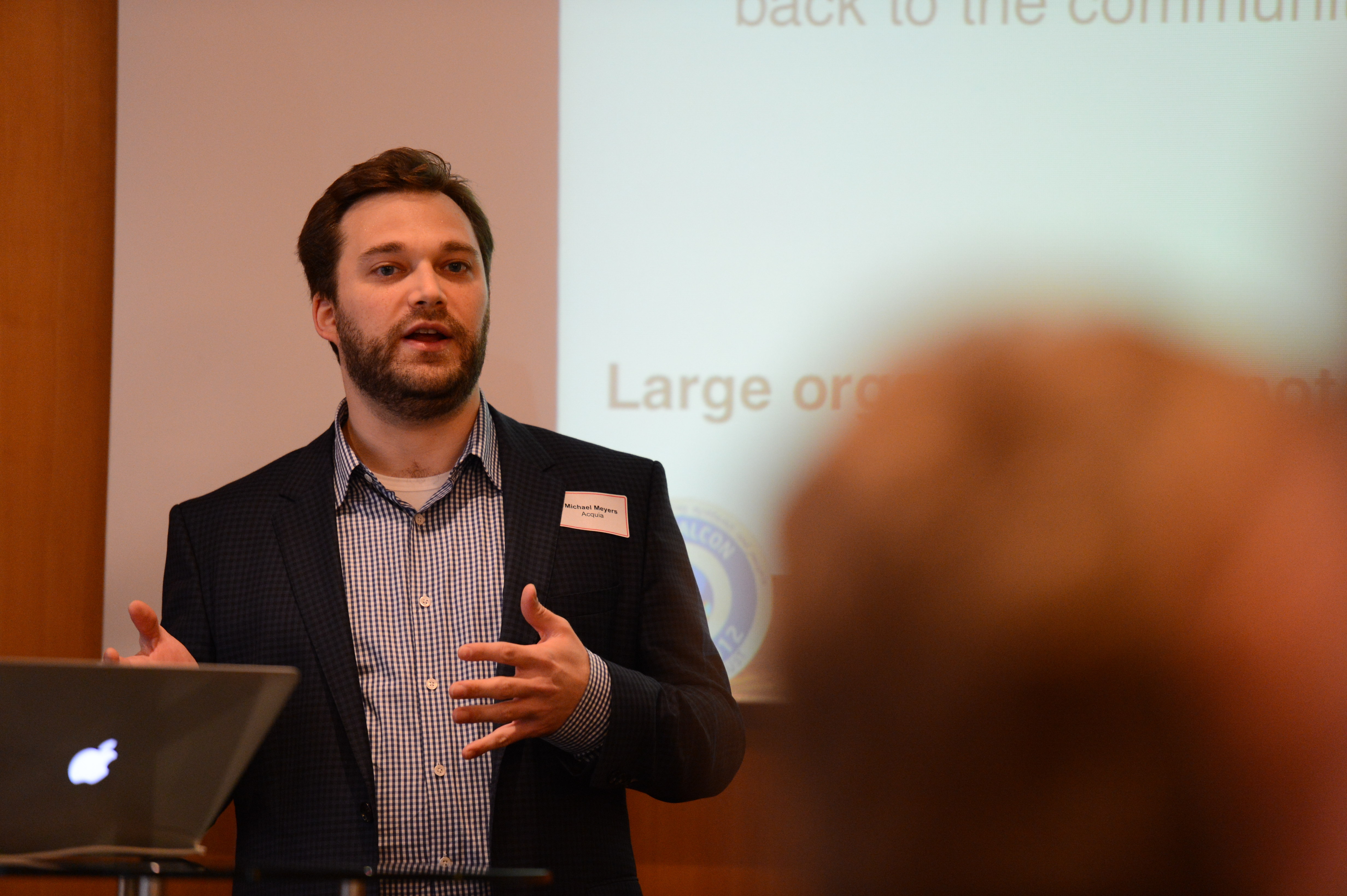 A speaker in a suit presents at DrupalCon Munich 2012, gesturing while addressing an audience with a projected slide.