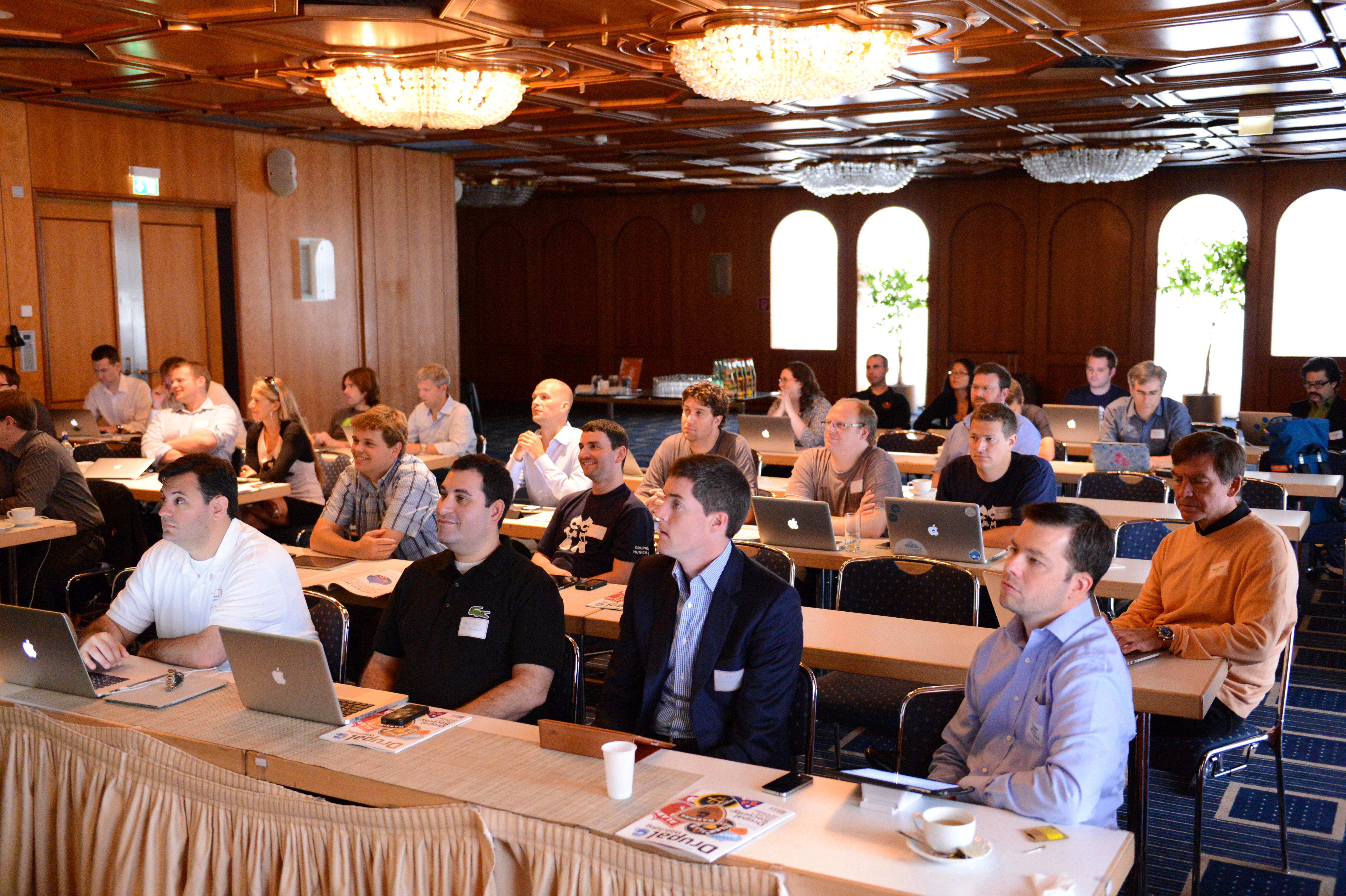 A large group of people attentively listening at a Drupal conference, with many using laptops in a conference room.
