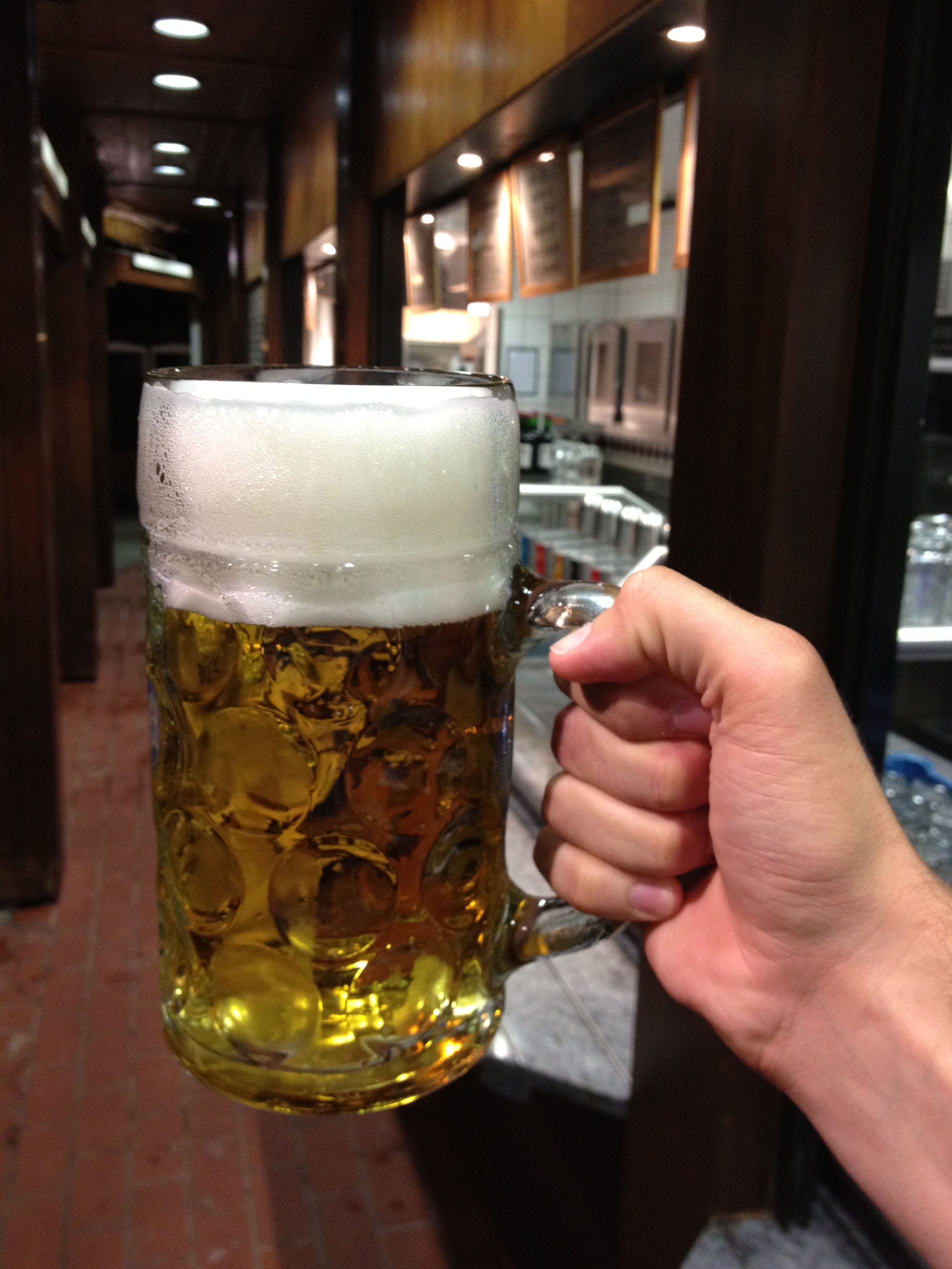 A person holds a large glass mug filled with beer, topped with foam, in a dimly lit bar.