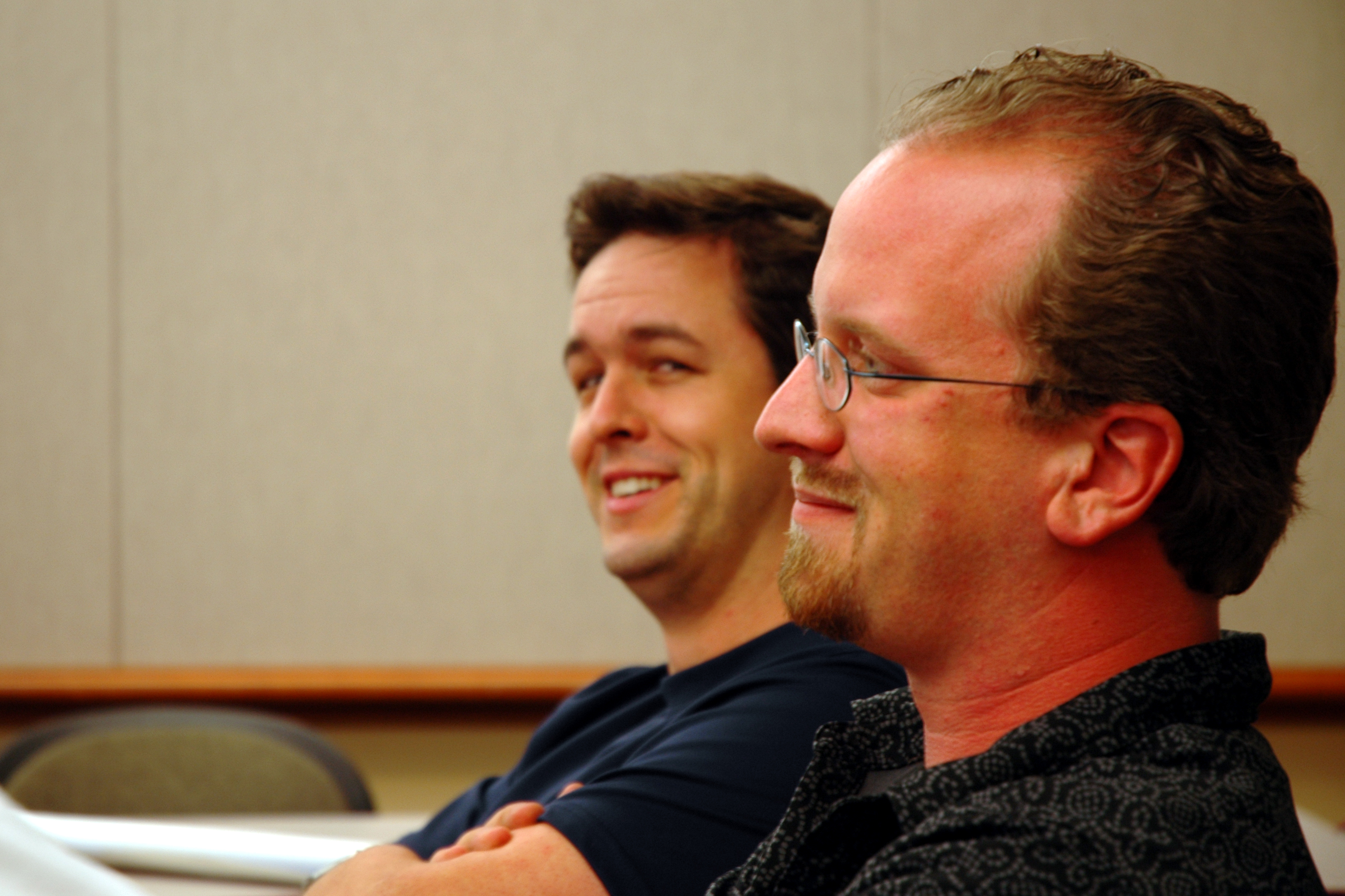 Two men sitting in a conference room, smiling and listening to a presentation at DrupalCon Portland 2005.