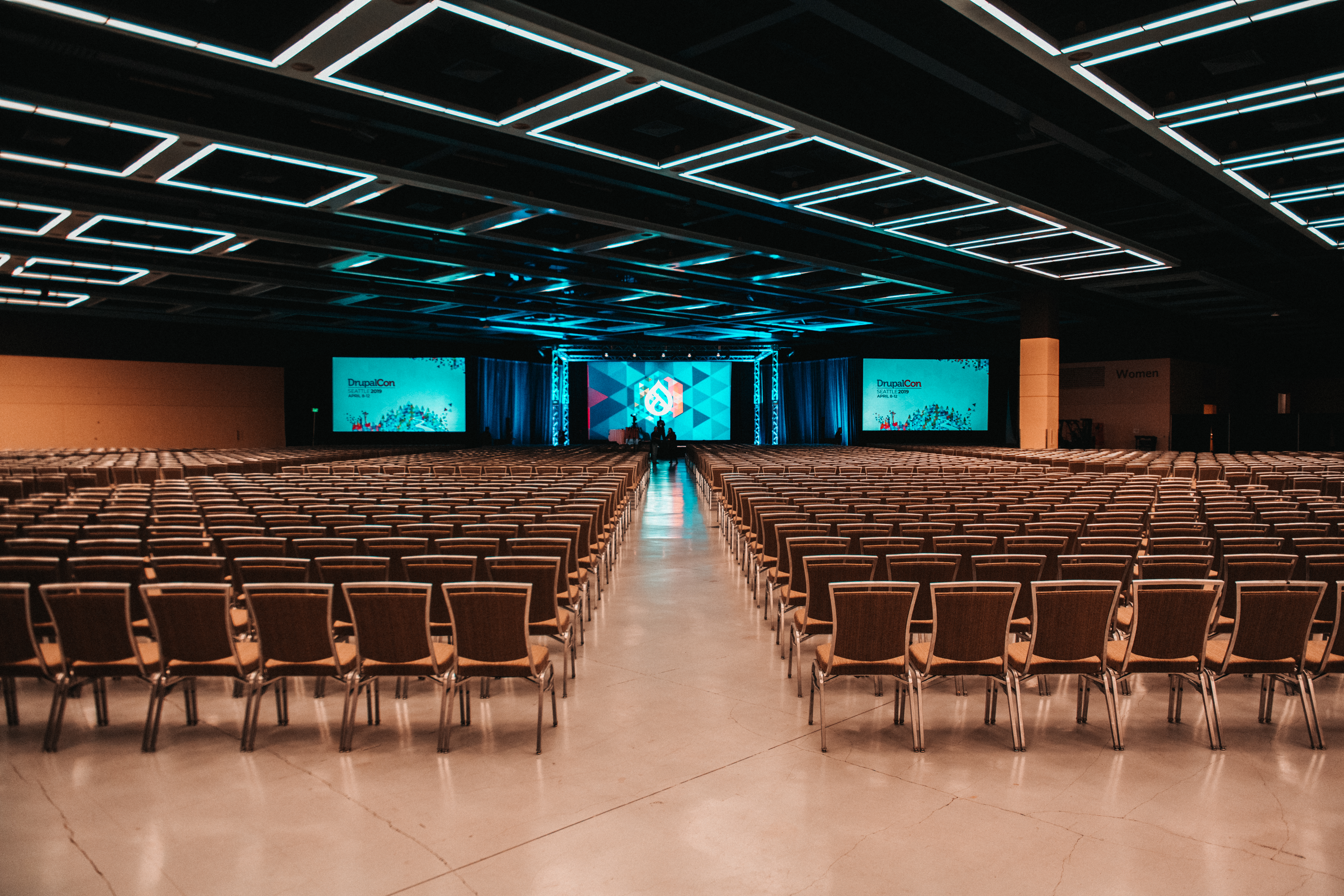 Large conference hall with rows of empty chairs facing a stage with big screens showing "DrupalCon" branding.