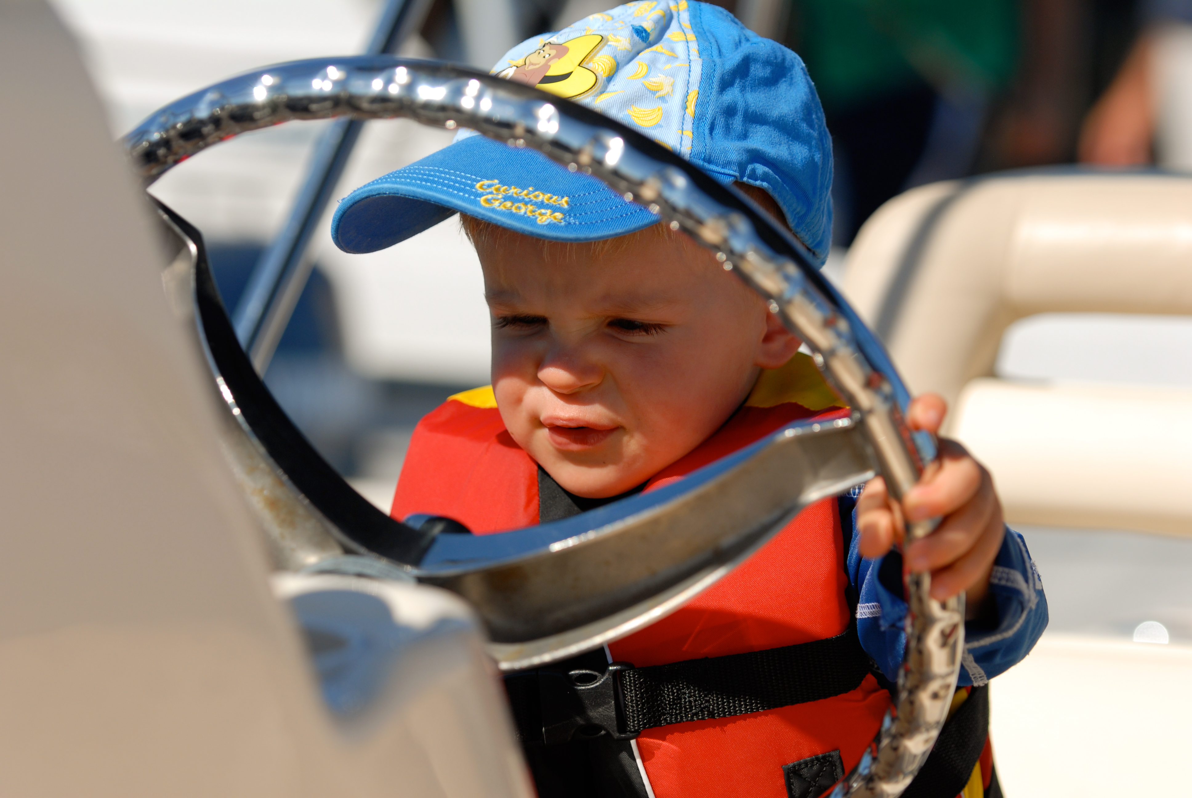 A young child wearing a life jacket and cap grips a boat's steering wheel with a focused expression.