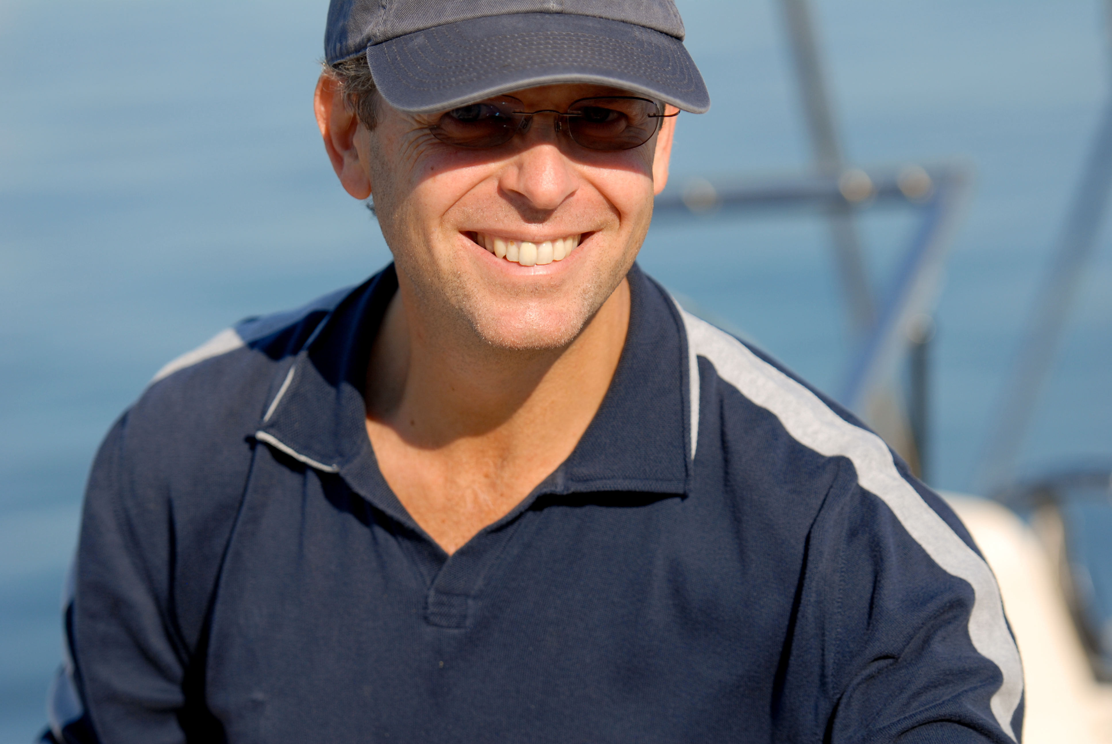 A man wearing sunglasses and a cap smiles while sitting on a boat in sunny weather.