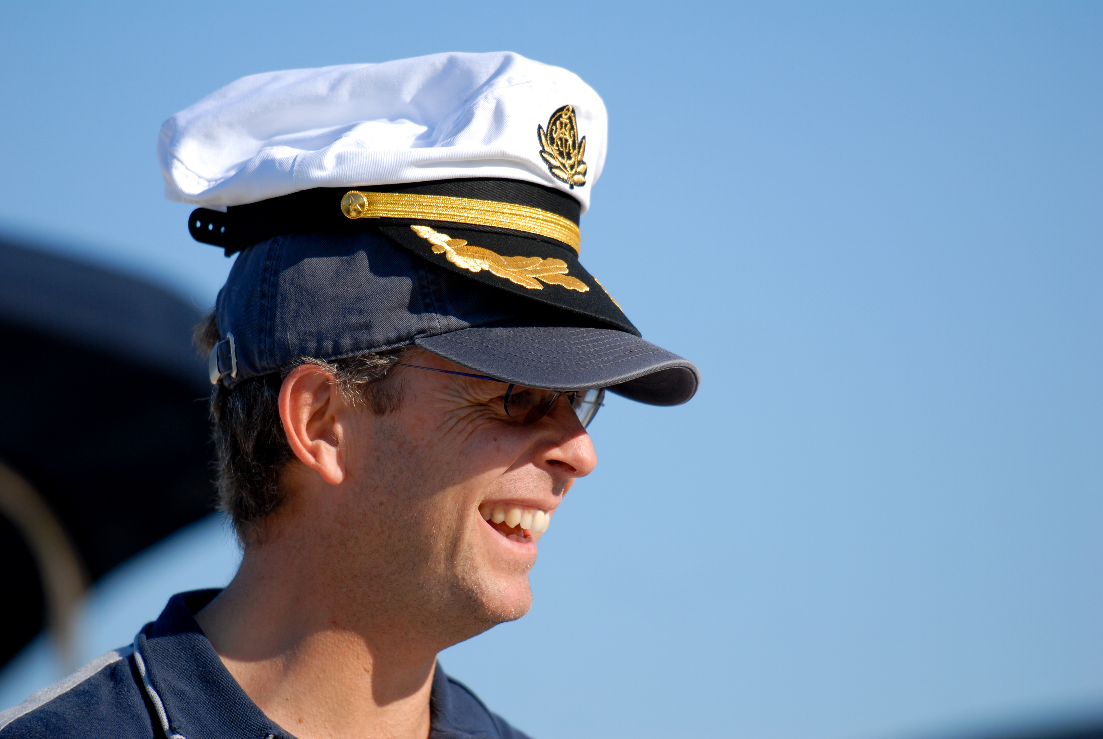 A man smiles while wearing two hats stacked on his head, including a captain's hat.