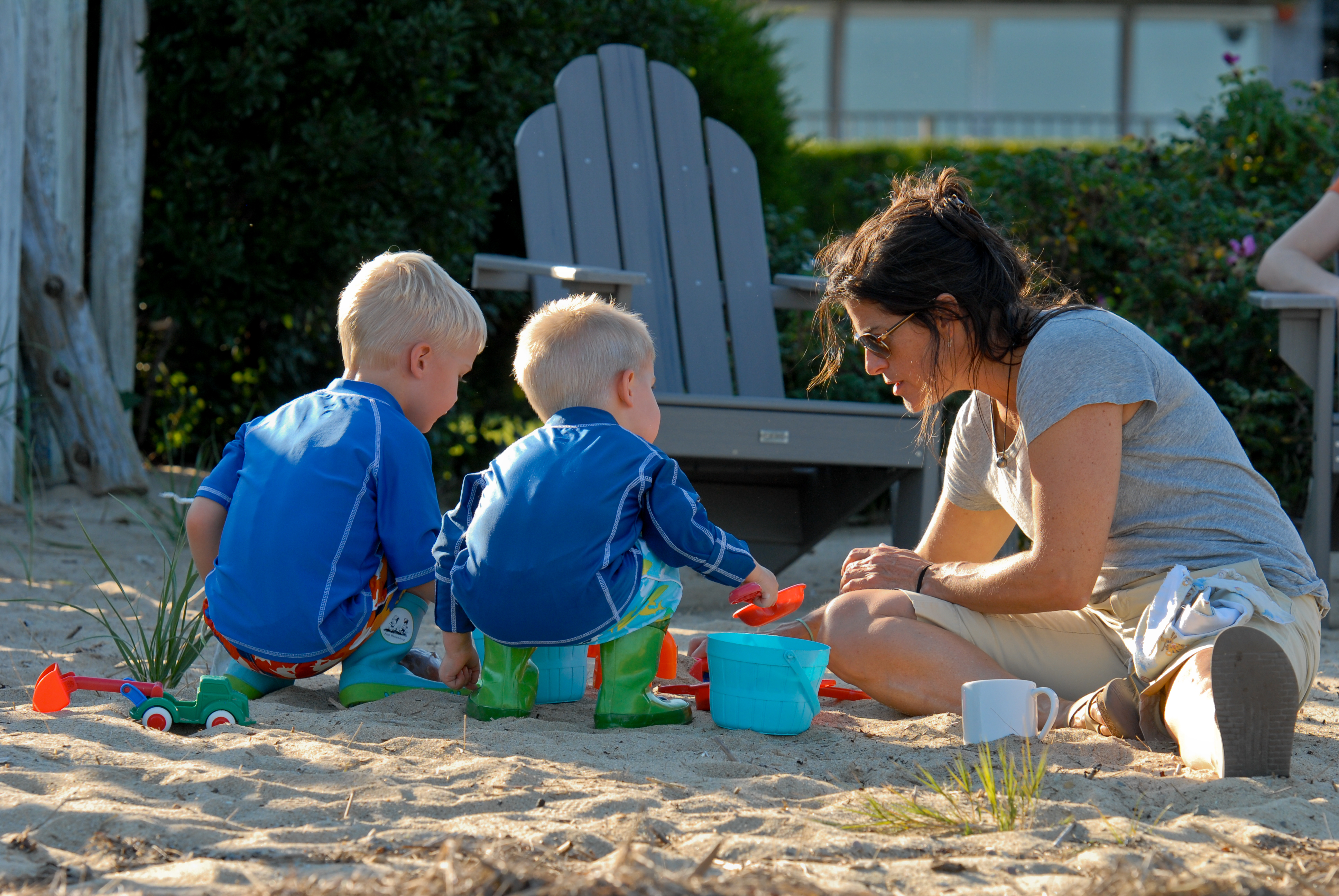 A woman and two young children play in the sand with buckets and shovels on a sunny day.