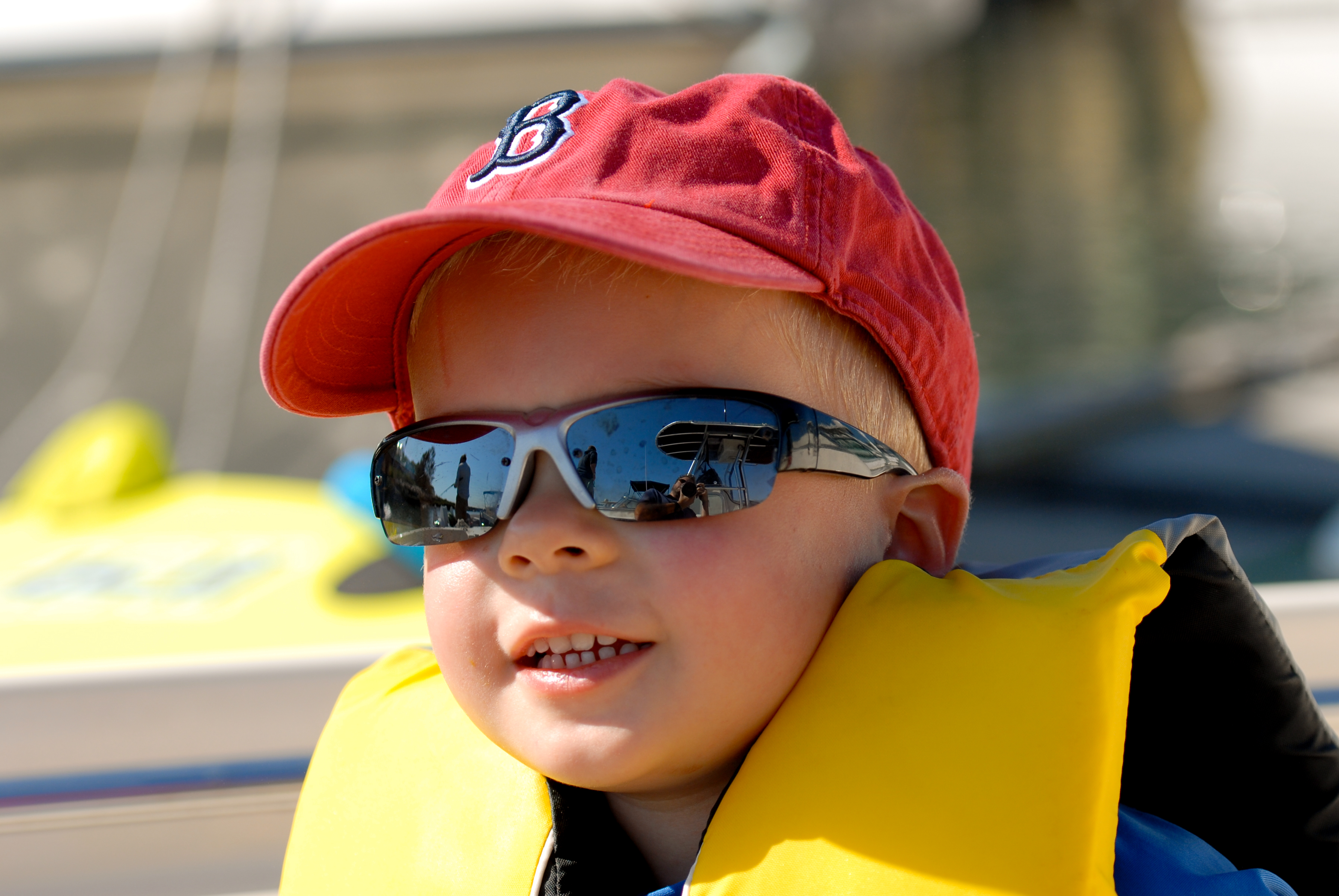 A young child wearing a red cap, sunglasses, and a life jacket smiles while sitting near the water.