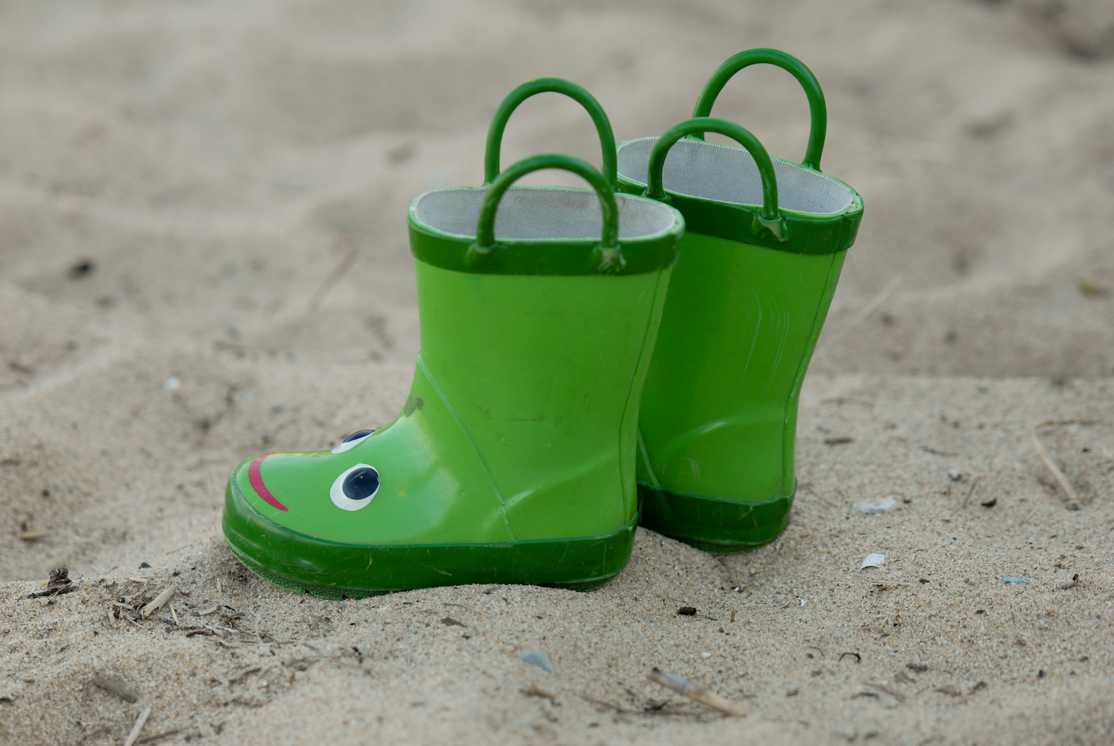 A pair of green children's rain boots with frog faces sit on sandy ground.