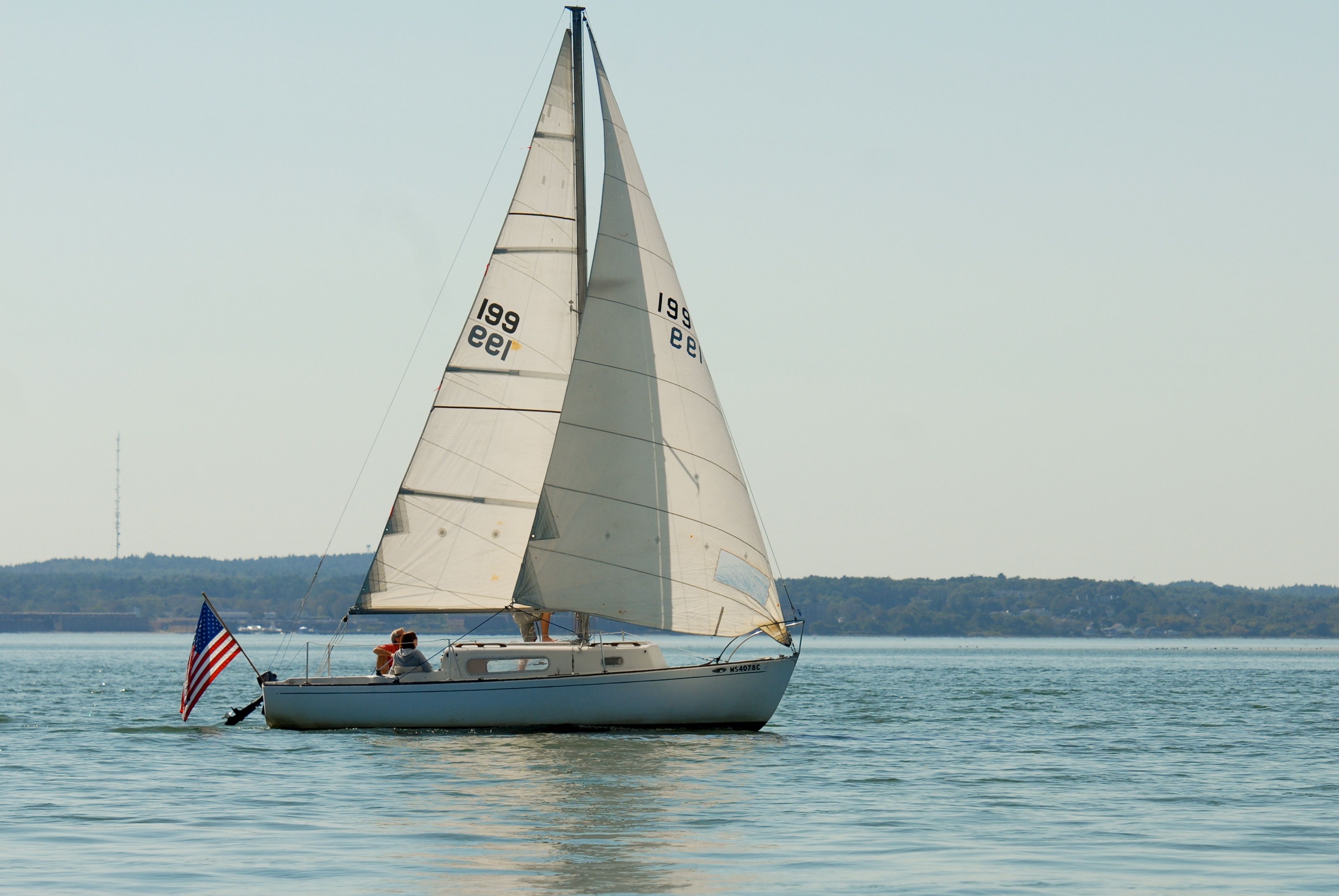A sailboat with two people on board moves across calm water, with an American flag at the stern.