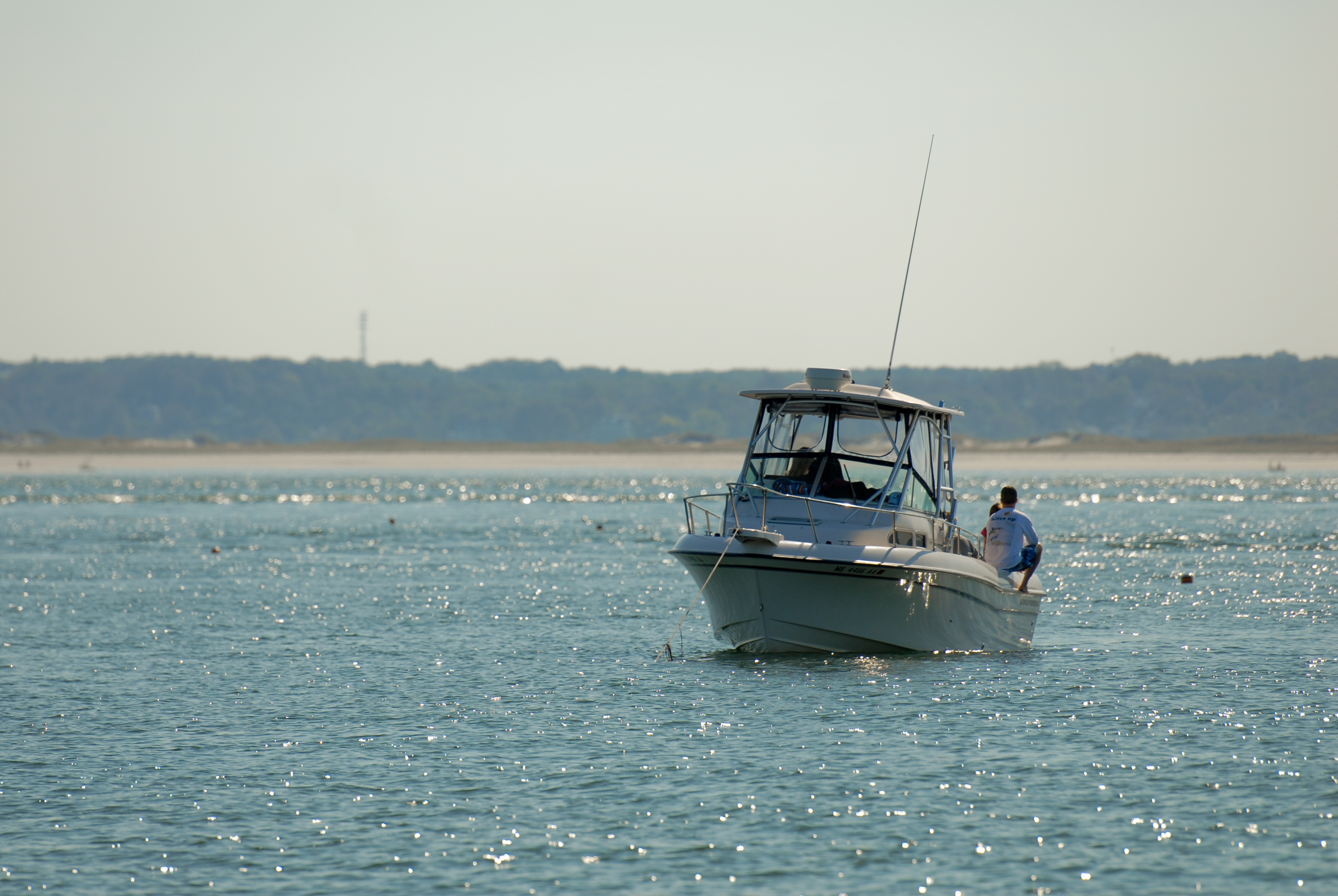 A person sits on the edge of a small motorboat floating on calm water near the shore.