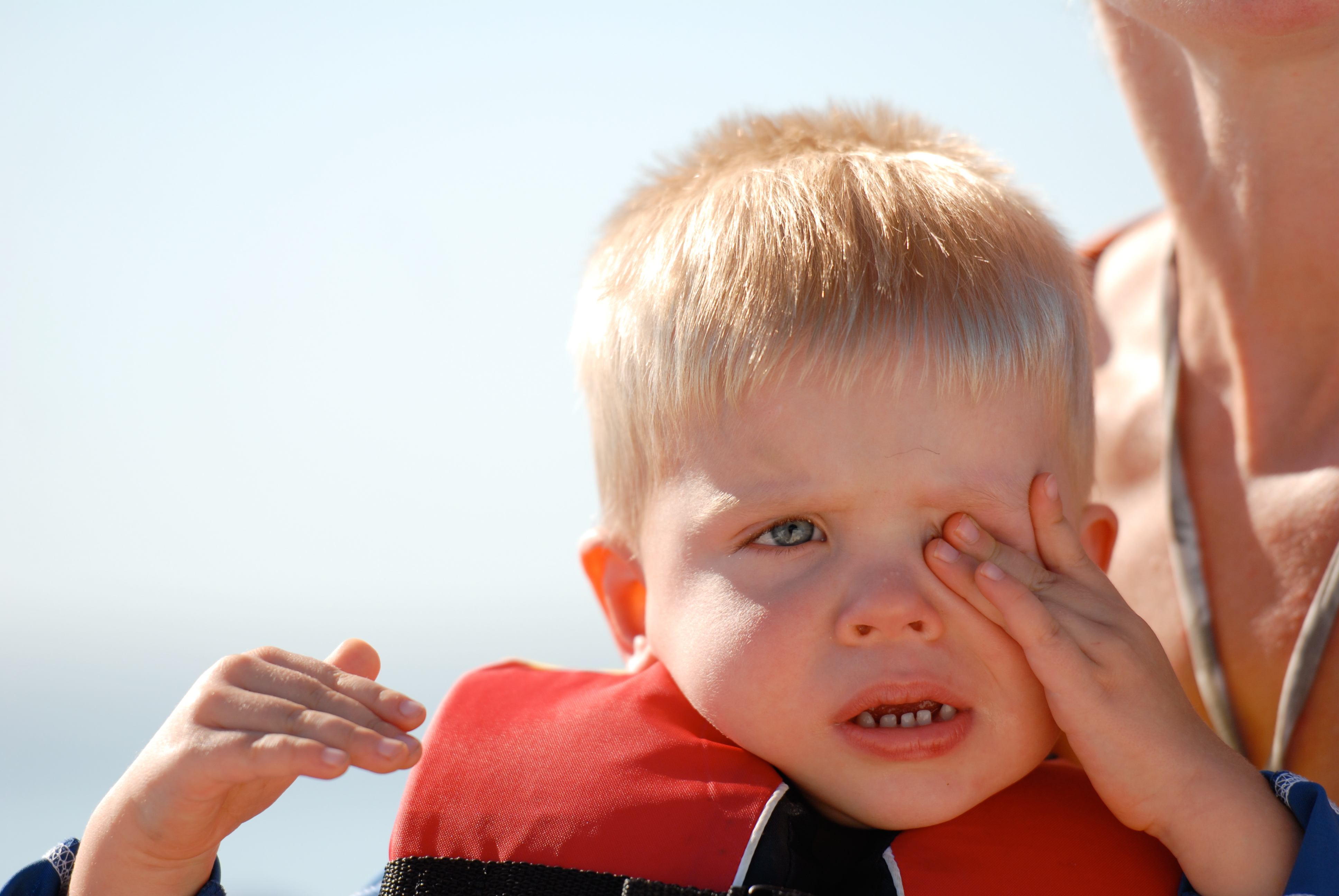 A young child in a life jacket rubs their eye while sitting with an adult in bright sunlight.