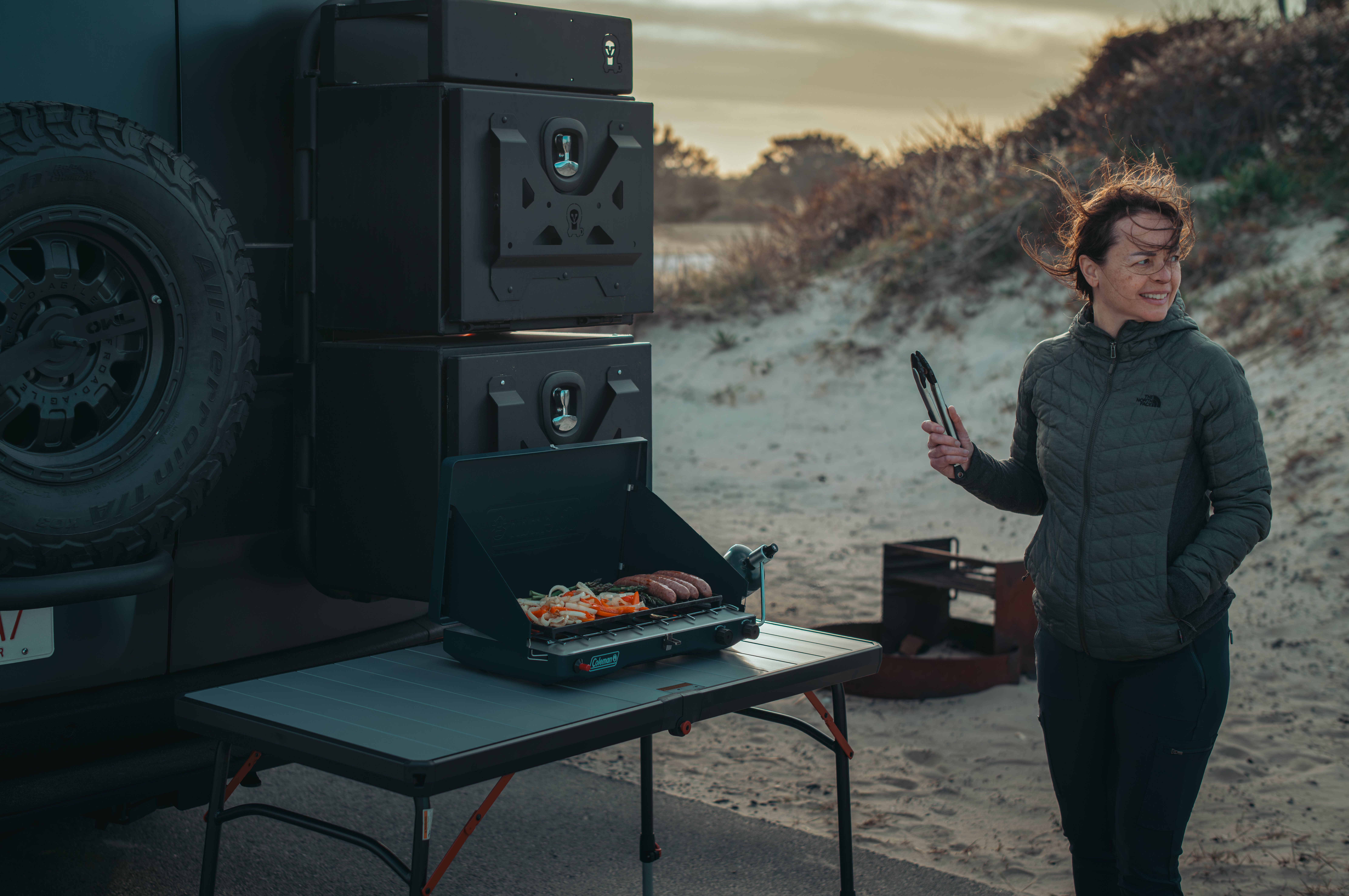 A woman behind a camper van grilling food near sandy dunes.