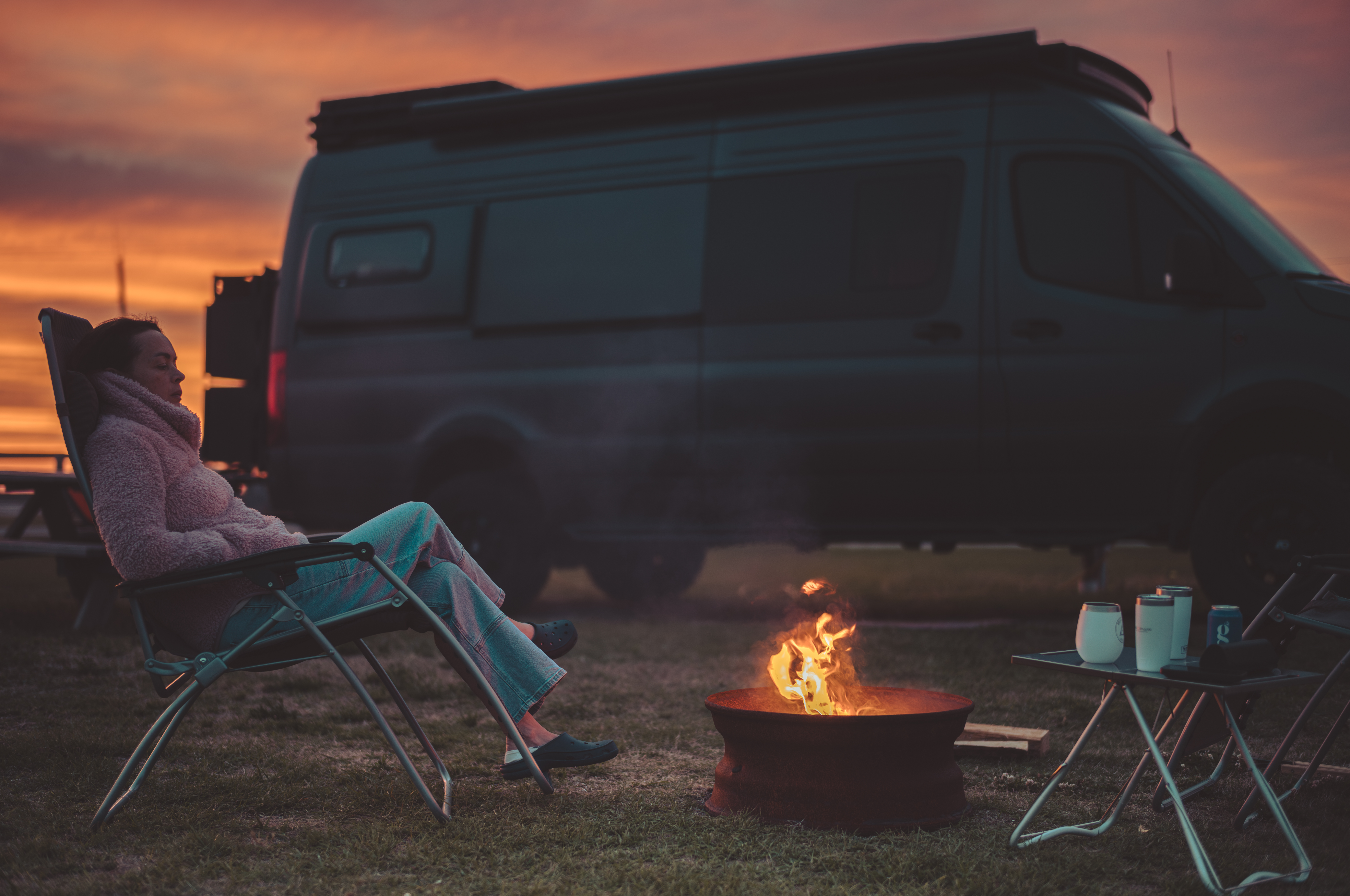 A woman reclines in a camp chair beside a fire pit at sunset with a camper van parked behind her.