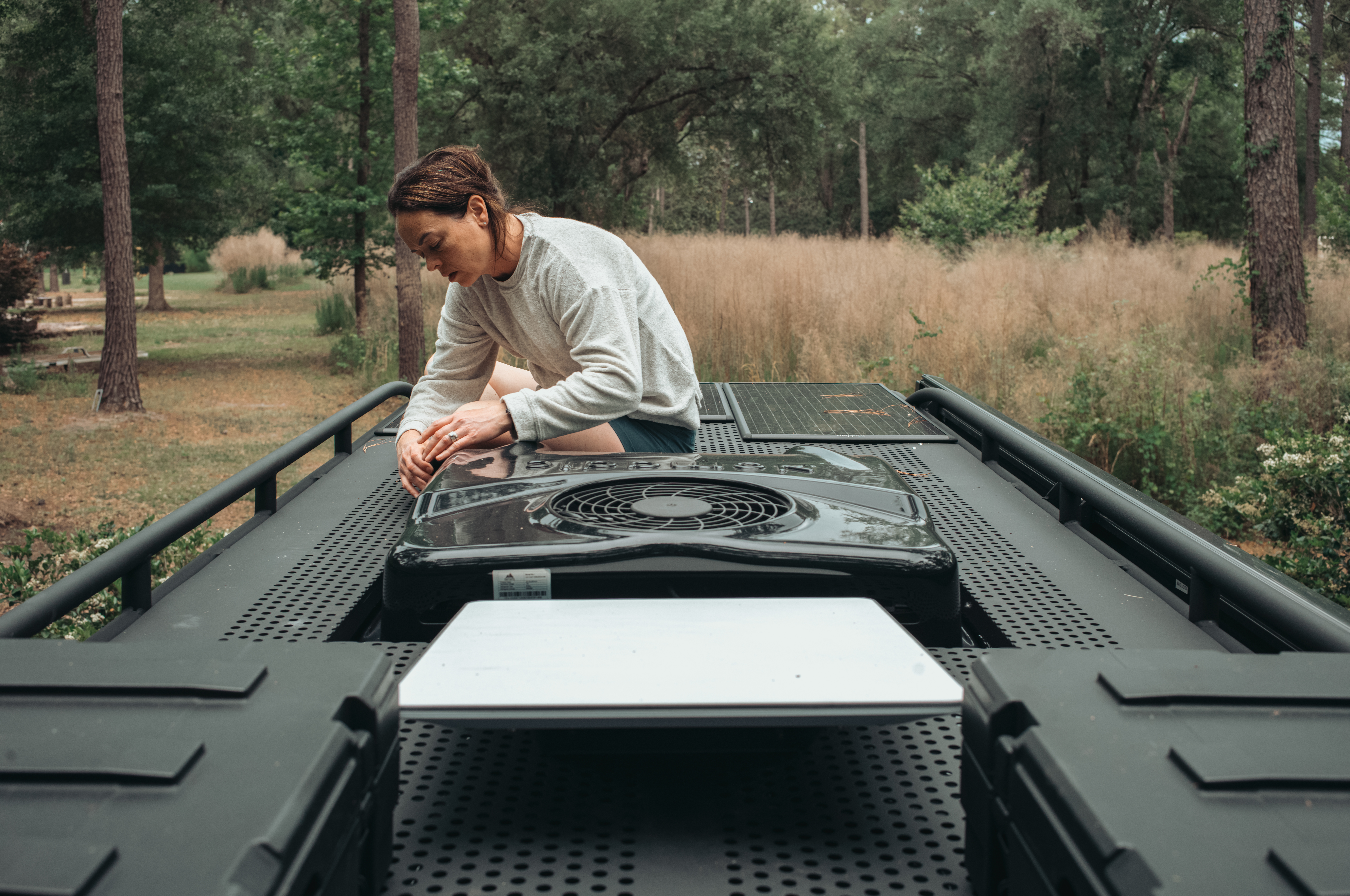A woman inspecting the A/C unit on the roof of a van at a wooded campsite.