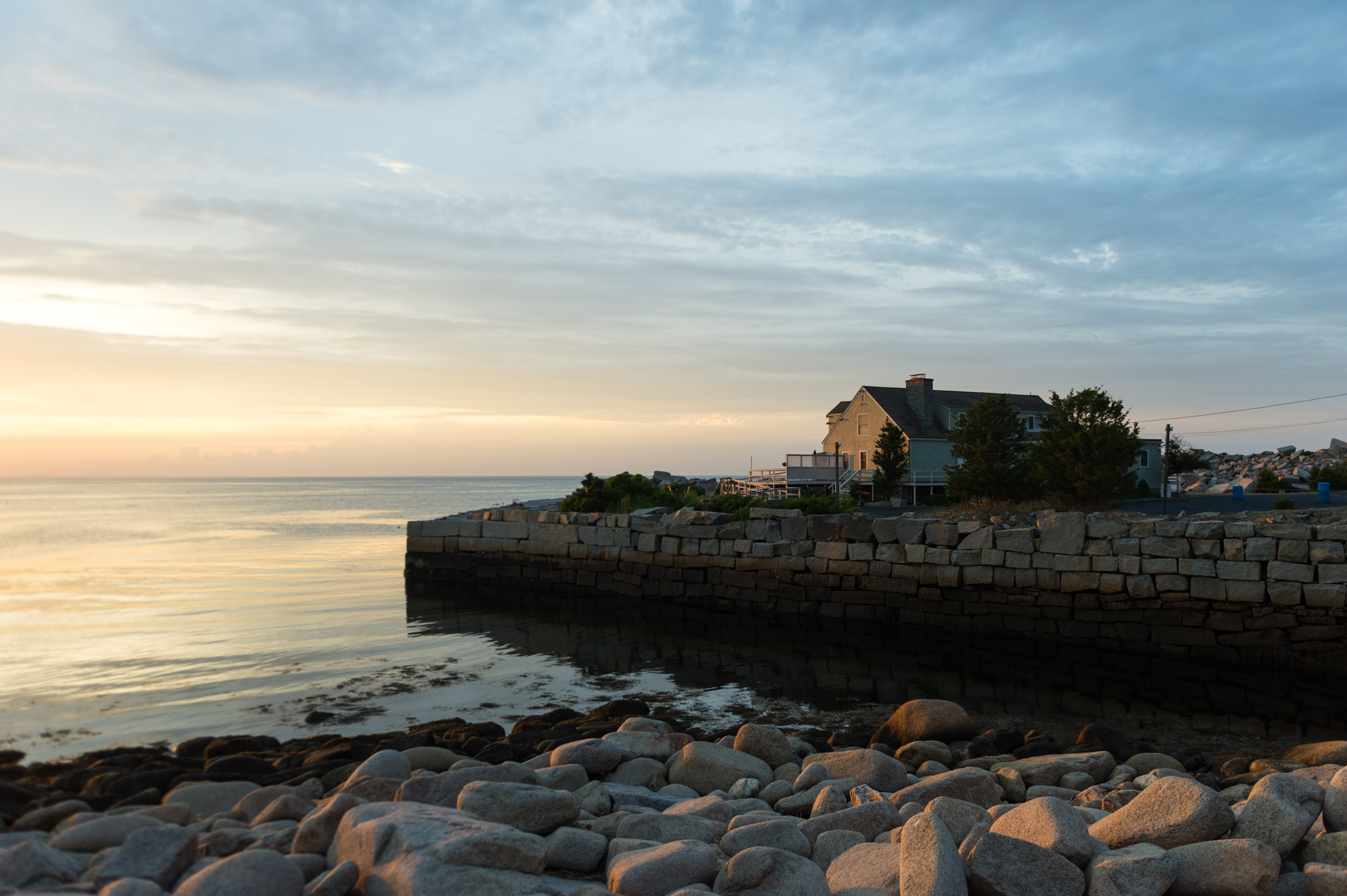 A coastal scene at sunset with a stone seawall, rocky shore, and a house overlooking calm waters.