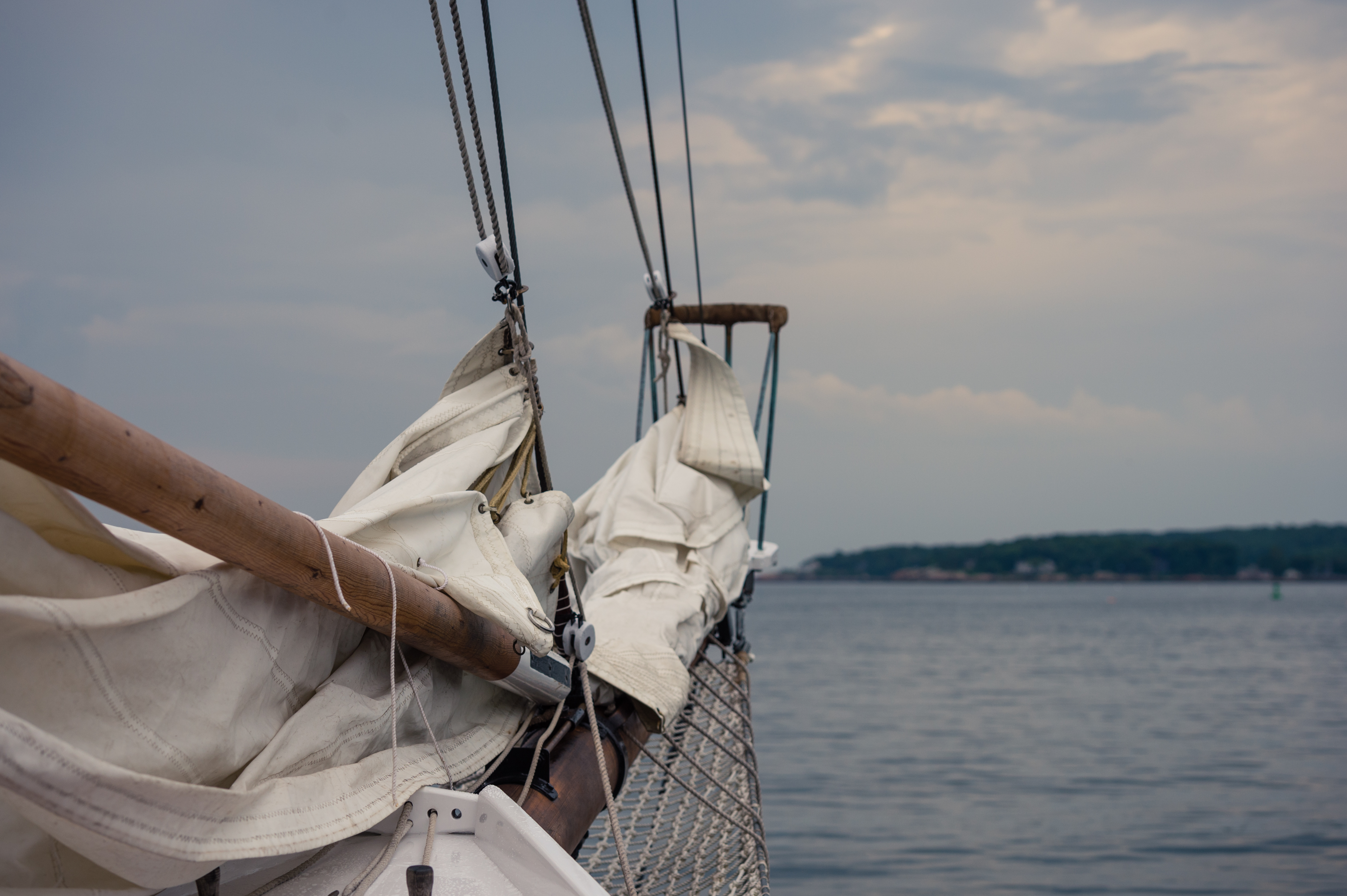 The front of a sailboat with furled sails, ropes, and rigging, floating on calm water under a cloudy sky.