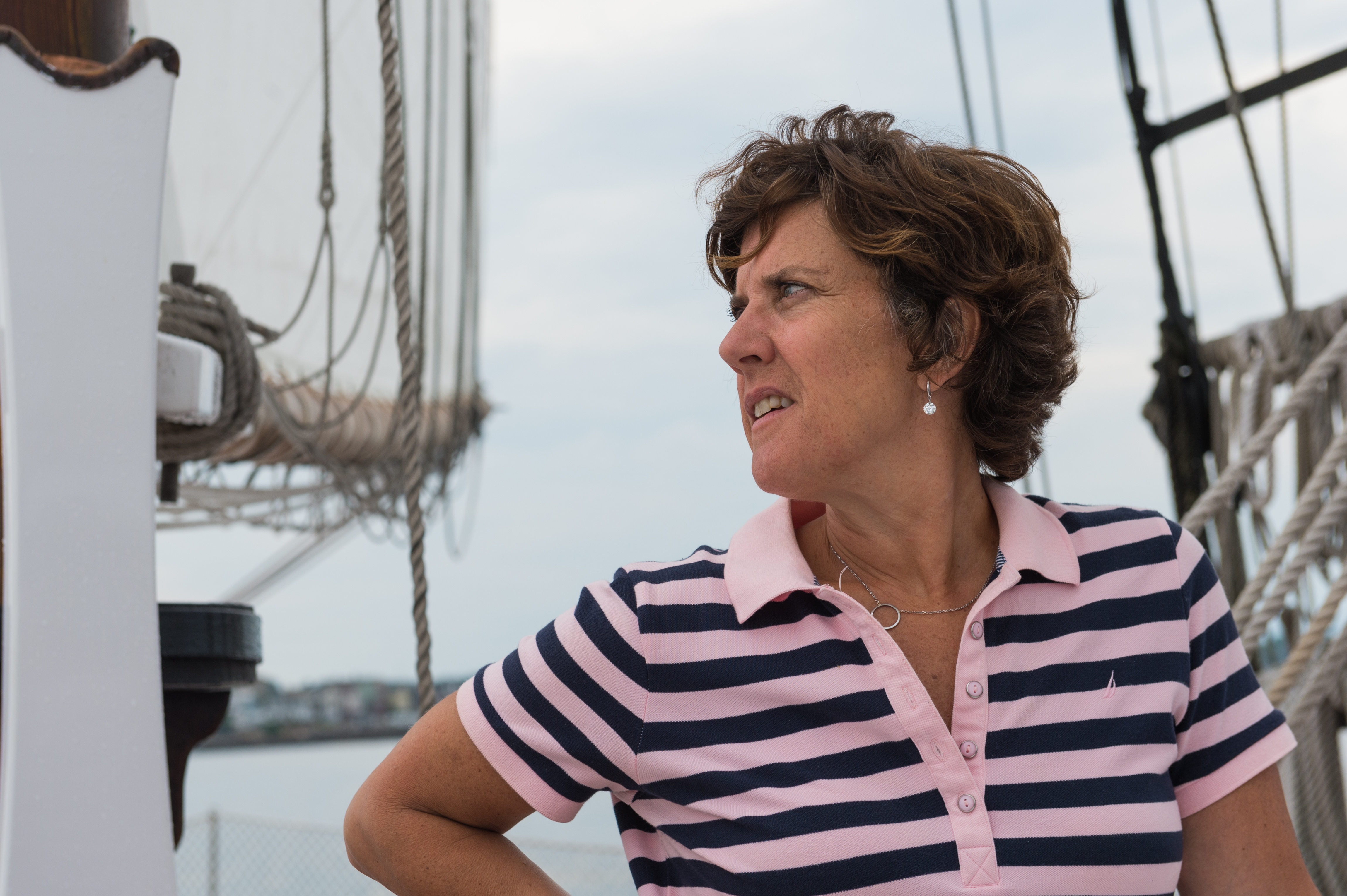 A woman in a striped polo shirt stands on a sailboat, looking to the side with a focused expression.