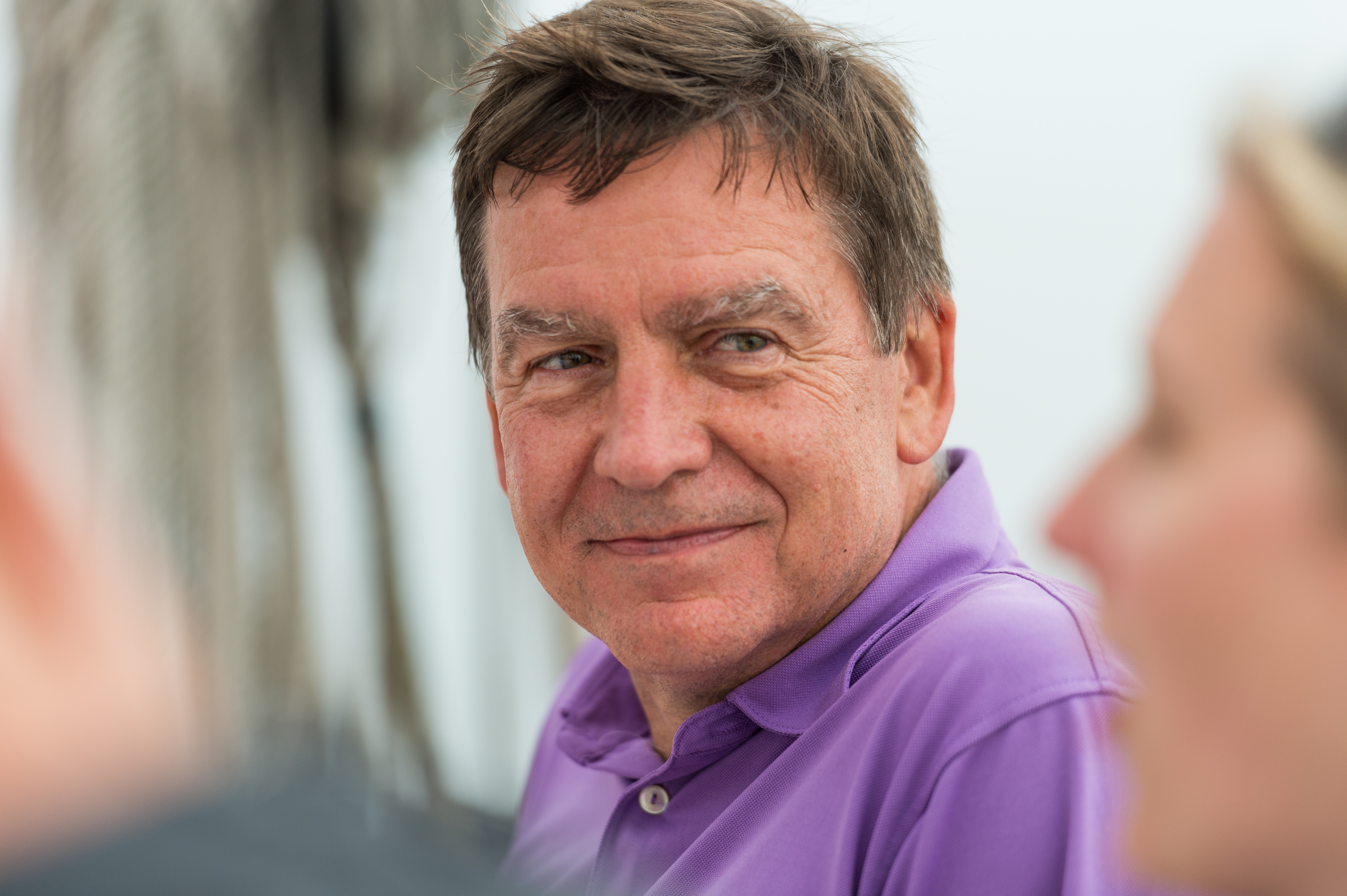 A man in a purple shirt smiles while talking to others on a sailing trip.