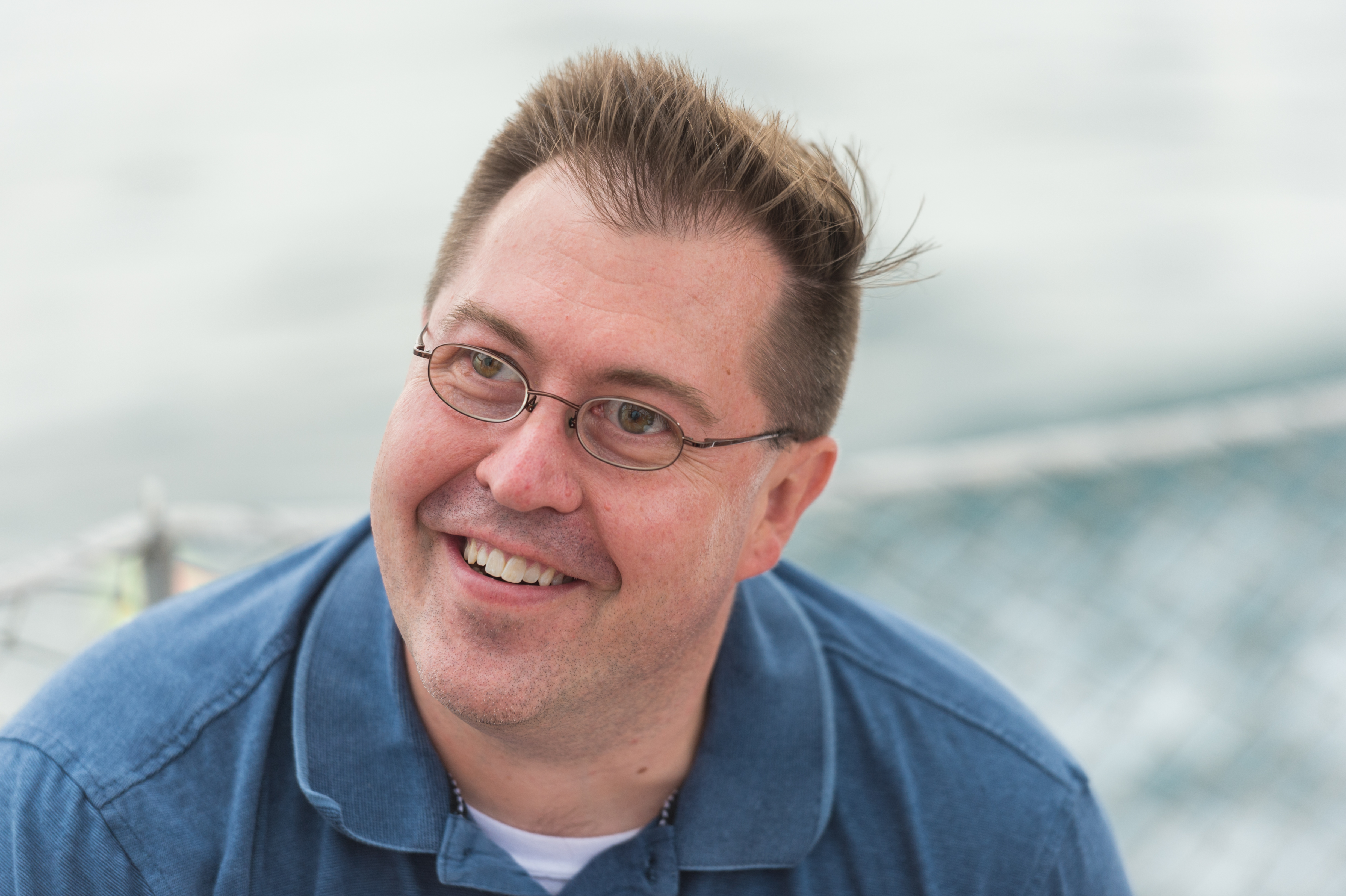 A smiling person wearing glasses and a blue shirt on a boat with water in the background.
