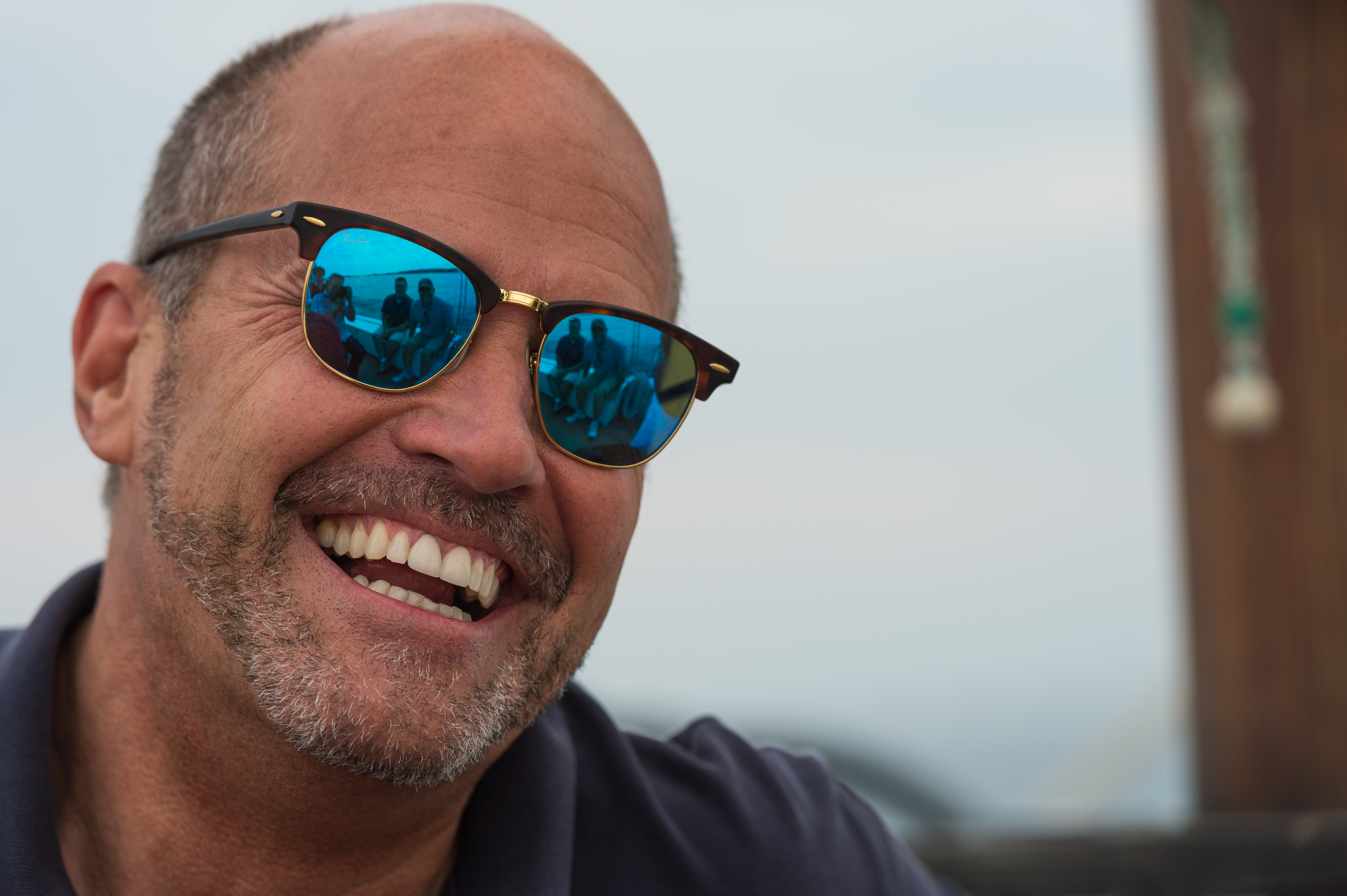 A smiling man wearing sunglasses reflects a group of people sitting together on a boat.