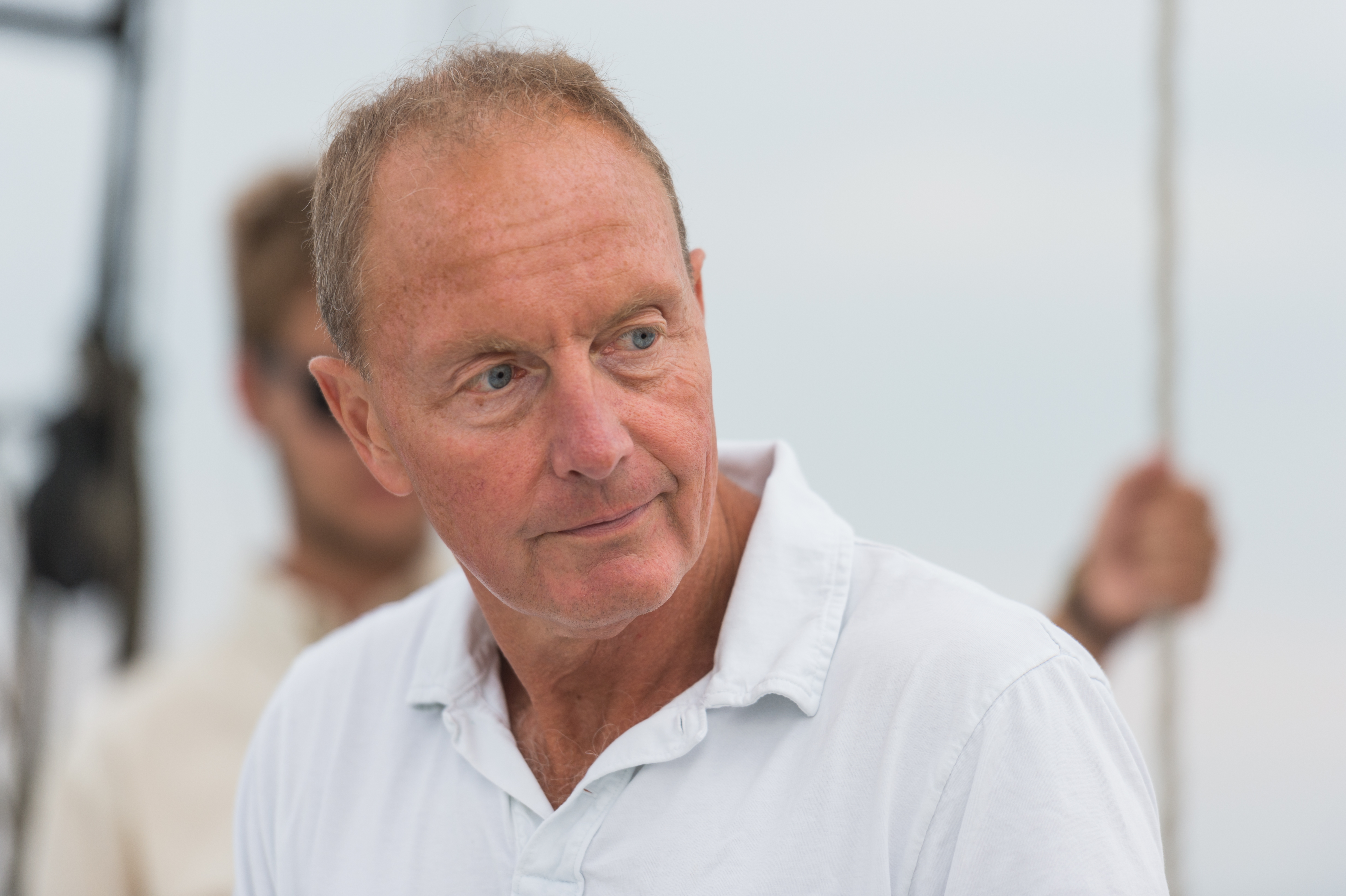 A man in a white polo shirt looks to the side while sailing, with another person holding a rope behind him.