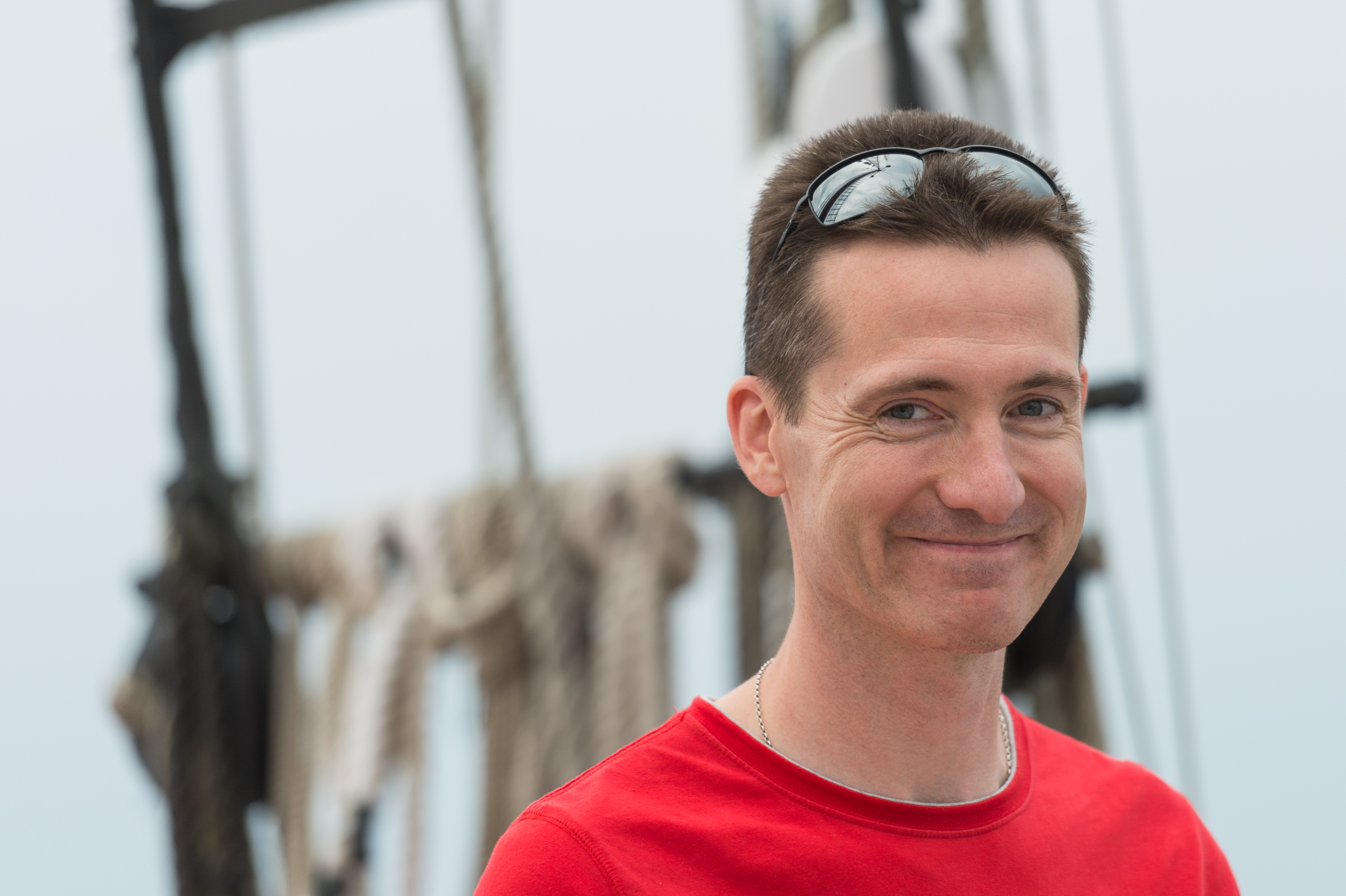 A man in a red shirt smiles while standing on a sailboat, with ropes and rigging in the background.