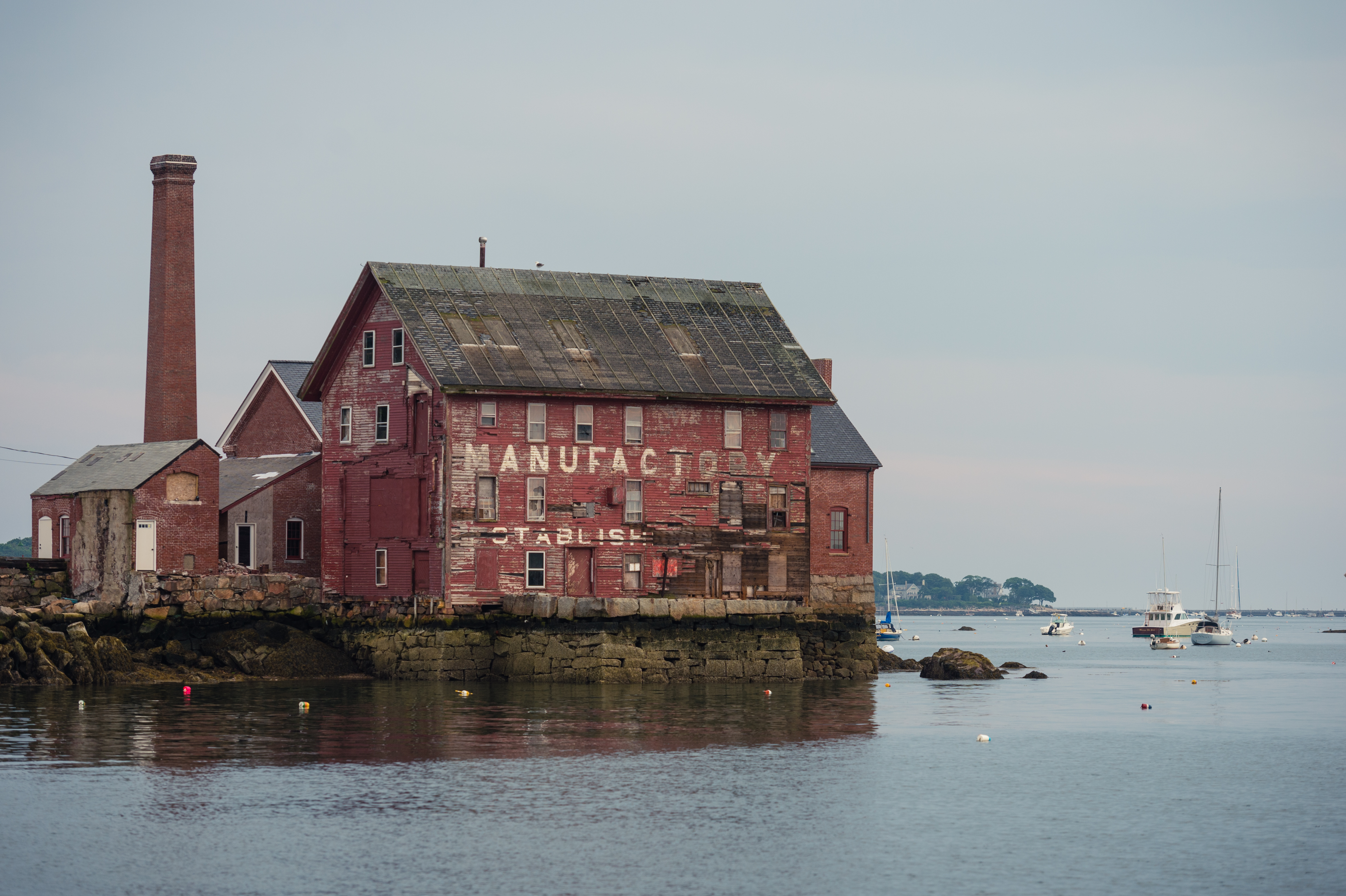 An old red brick building by the water with sailboats and motorboats anchored nearby.