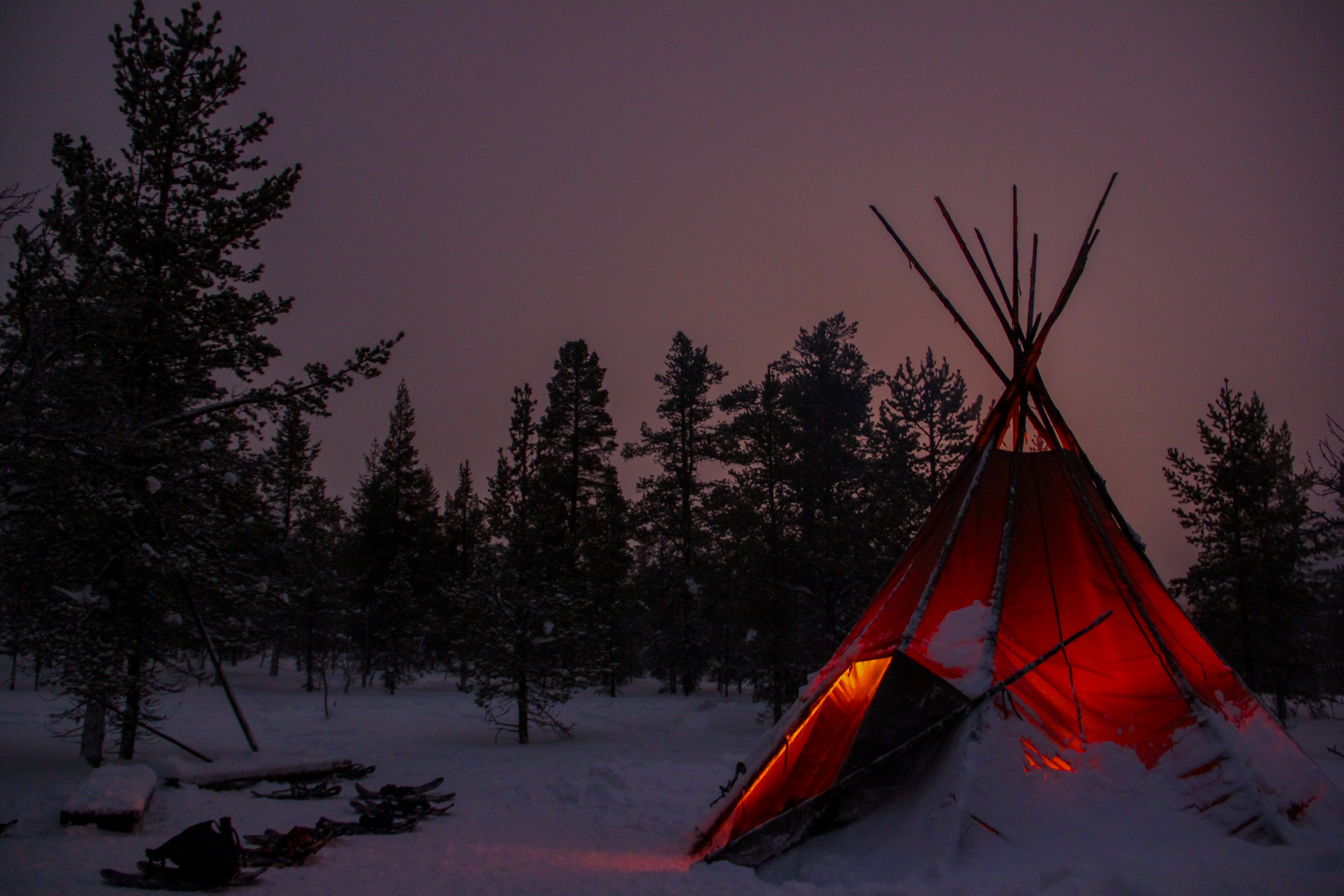 A red teepee with a glowing interior stands in a snowy forest at night, surrounded by trees and scattered gear.
